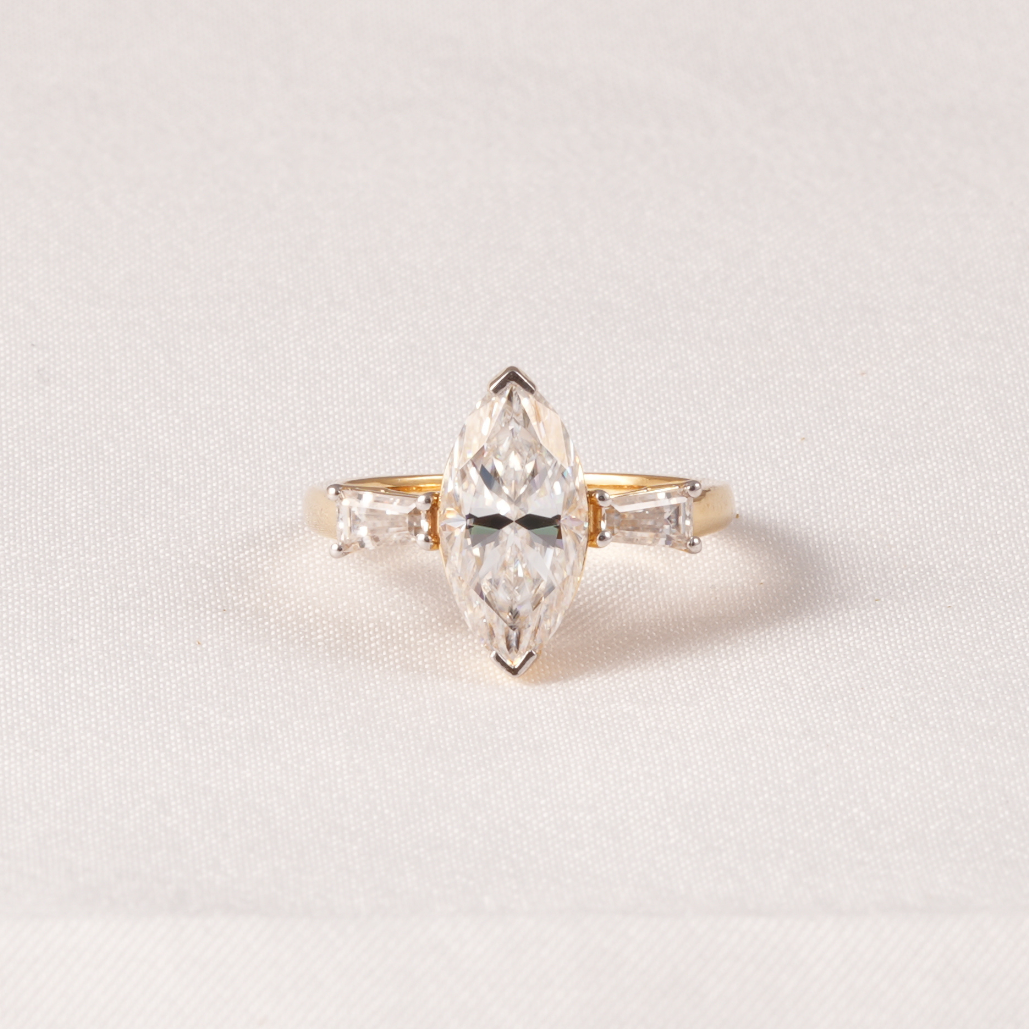 A yellow gold ring featuring a marquise-cut clear center diamond flanked by two baguette-cut side diamonds, set against a soft, light beige textured fabric background.