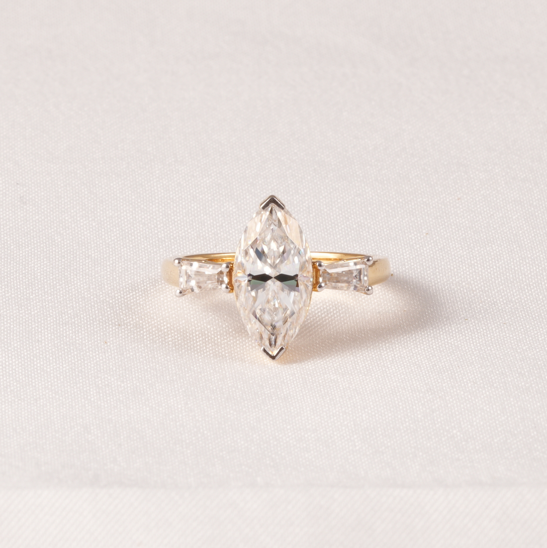 A yellow gold ring featuring a marquise-cut clear center diamond flanked by two baguette-cut side diamonds, set against a soft, light beige textured fabric background.