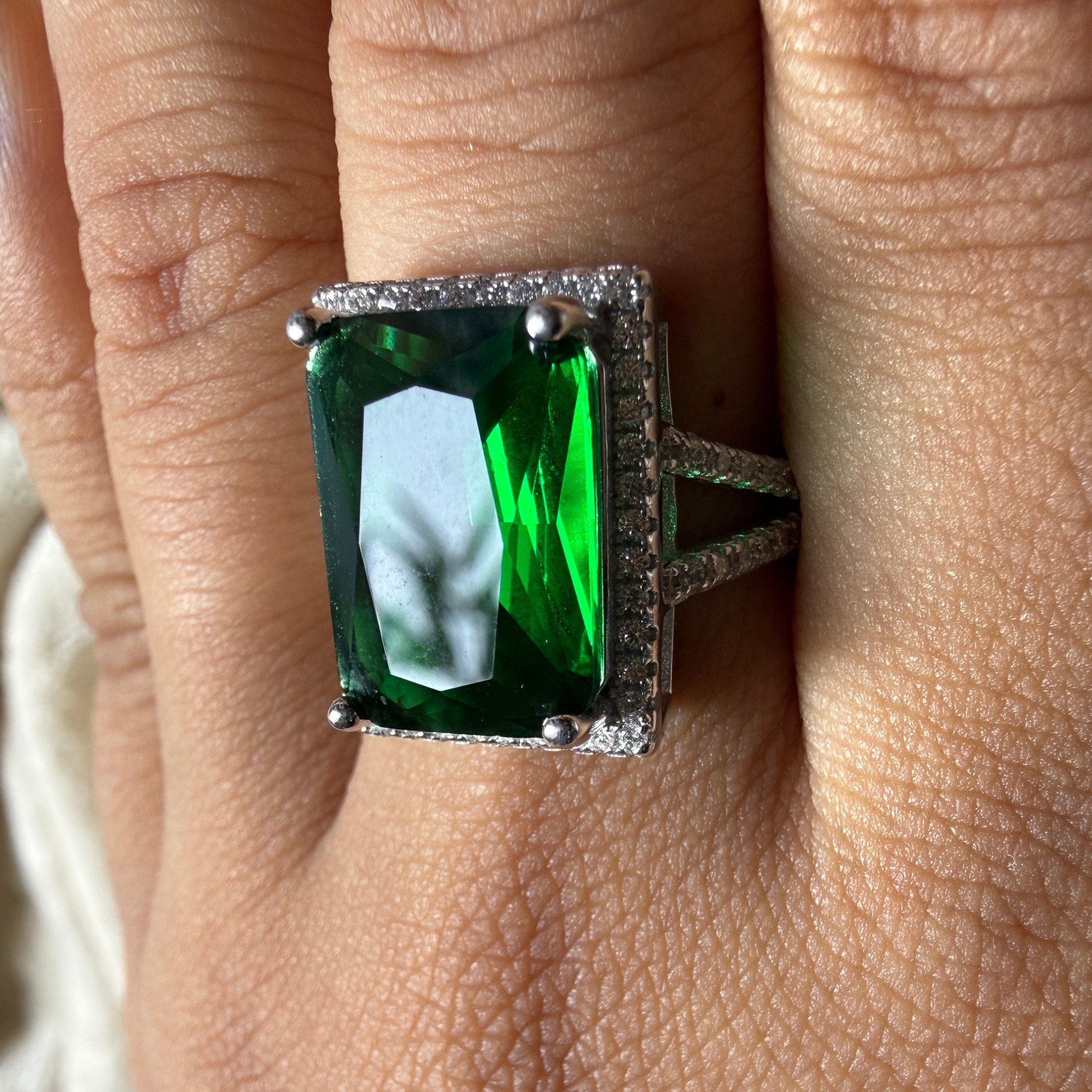A rectangular emerald-cut green gemstone ring with a silver-toned halo and band, shown worn on a person's finger.