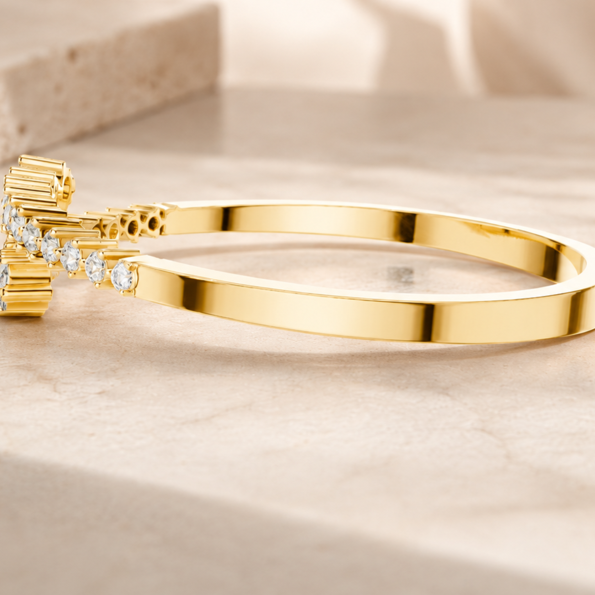 A polished yellow gold hinged bangle bracelet with a decorative clasp featuring small clear gemstones, resting on a light beige stone surface.