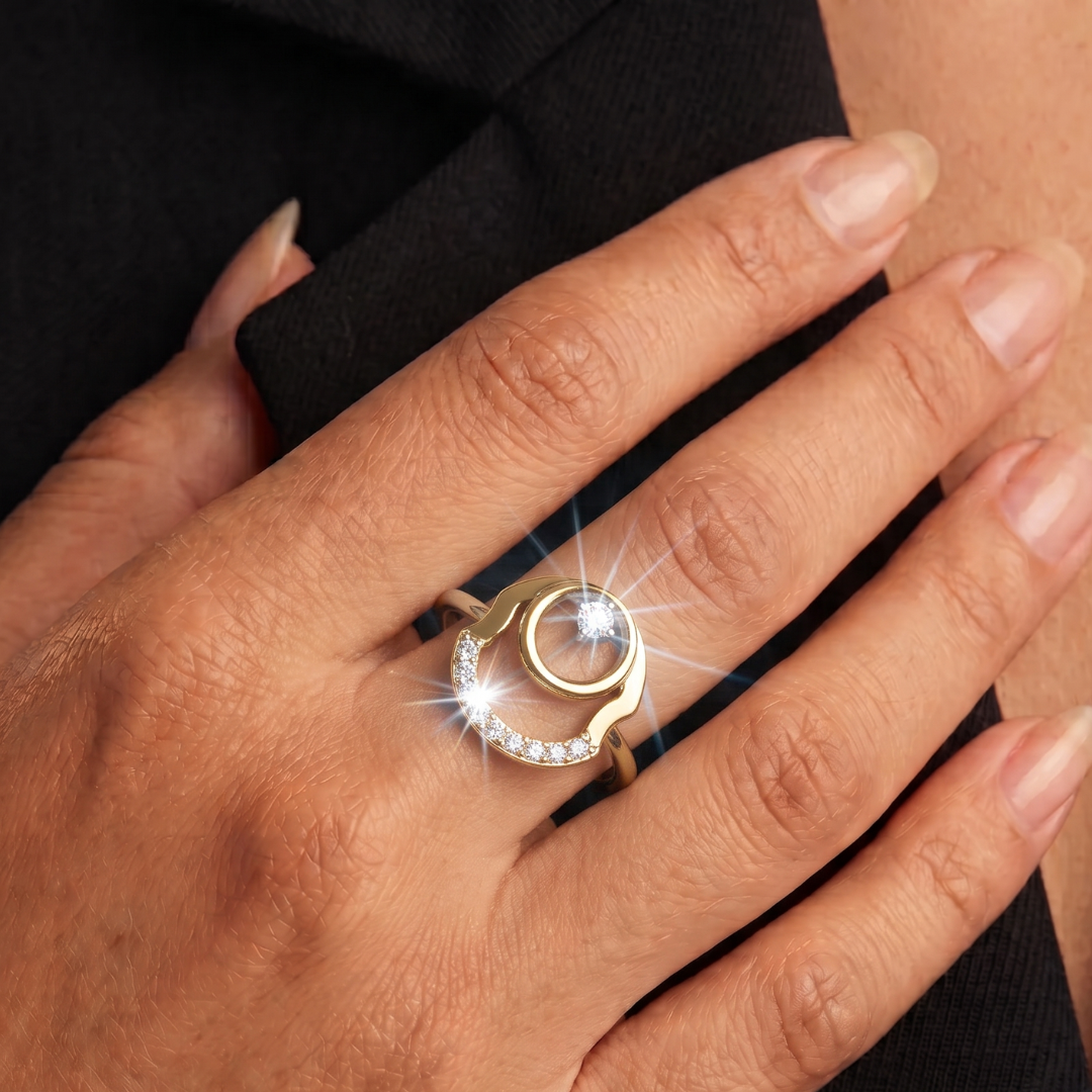 A gold ring with a central clear gemstone and a curved halo of small clear stones, shown worn on a person's finger against a black sleeve.