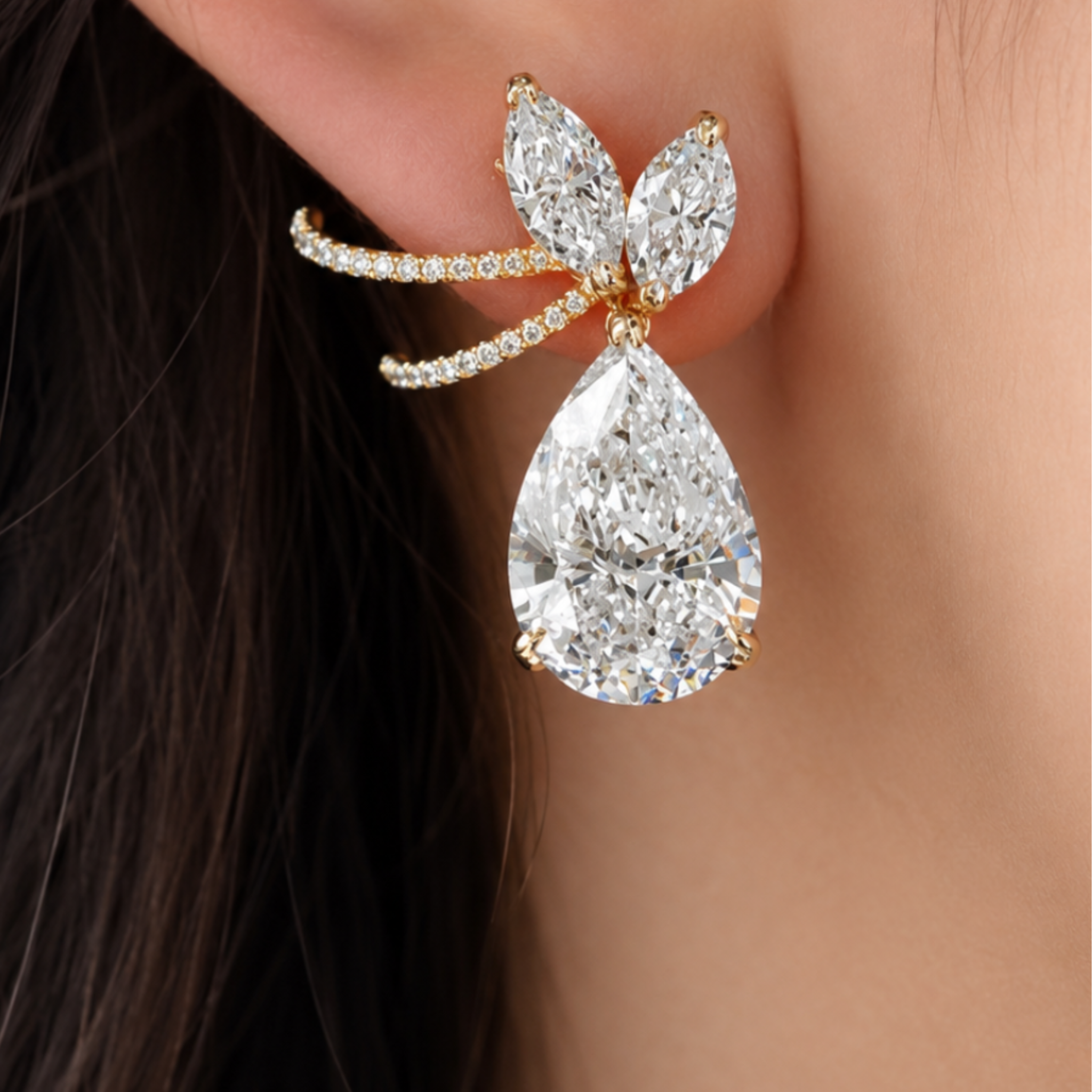 A close-up of a gold ear cuff earring worn on a person's ear, featuring a large pear-cut clear gemstone and smaller marquise-cut stones with tiny pavé accents.