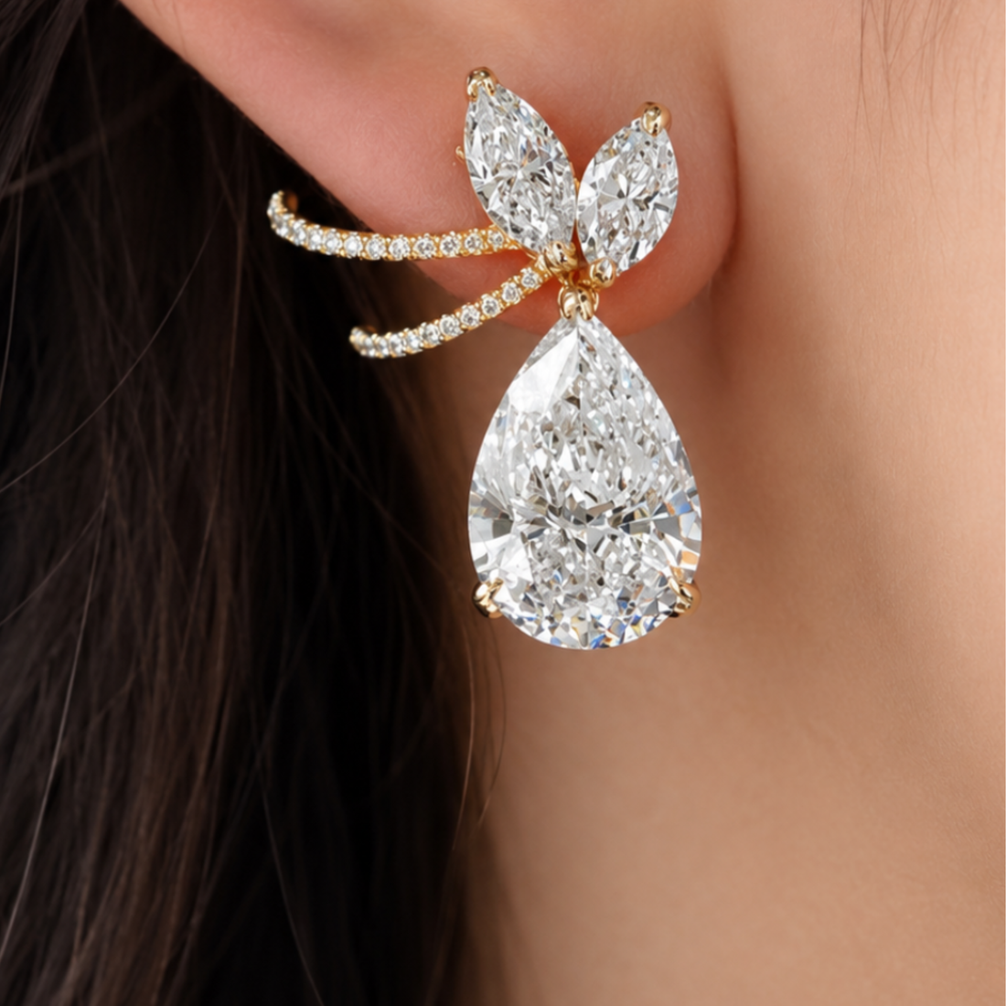 A close-up of a gold ear cuff earring worn on a person's ear, featuring a large pear-cut clear gemstone and smaller marquise-cut stones with tiny pavé accents.