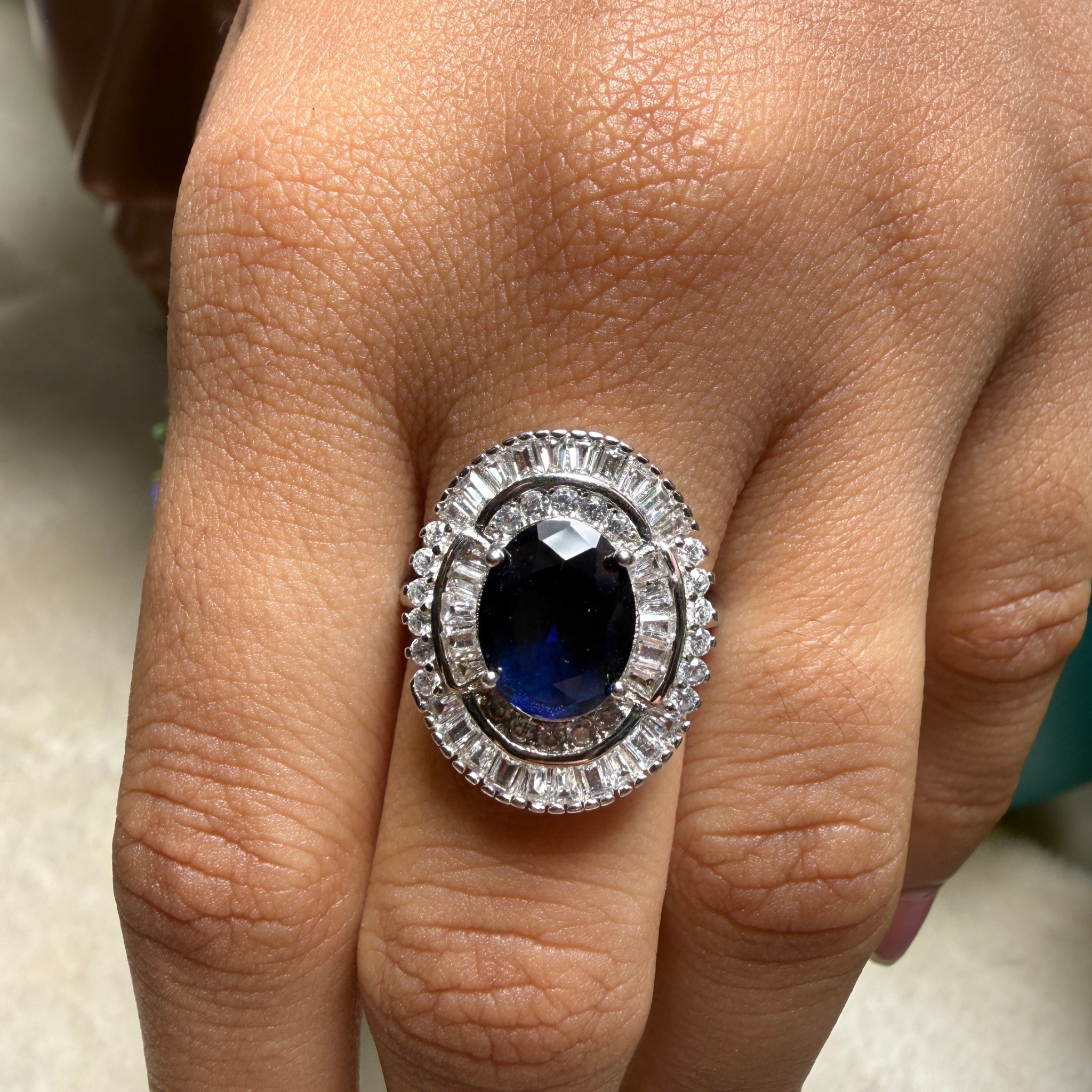 A close-up of a hand wearing a large oval ring with a deep blue center stone and a halo of clear stones in a silver-toned setting.