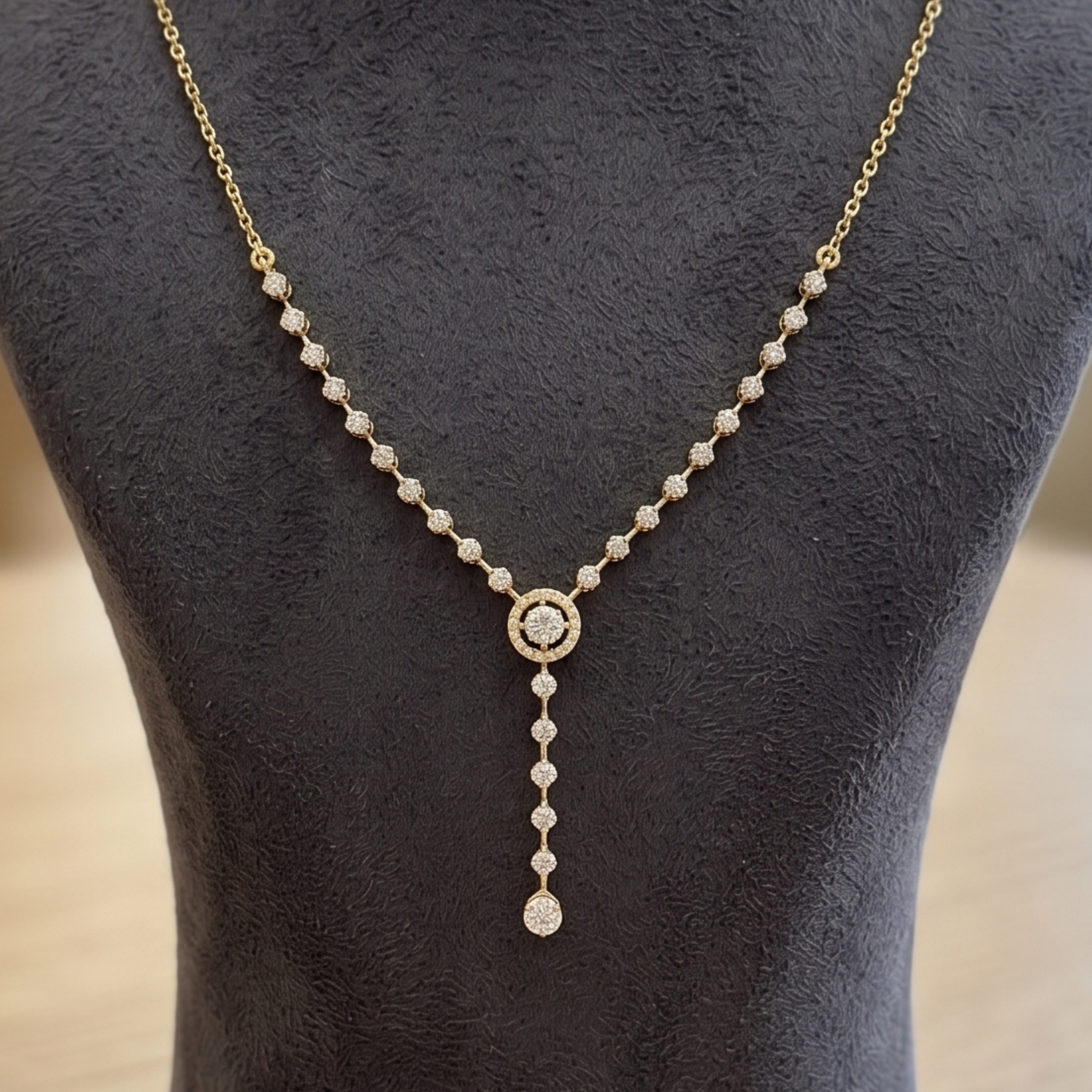 A delicate gold Y-necklace with clear pavé-set round stones, featuring a central round stone surrounded by a halo and a teardrop-shaped drop at the bottom, displayed on a dark textured jewelry bust.