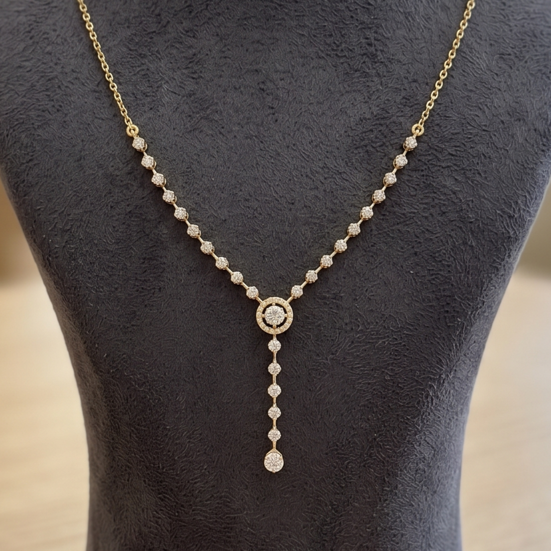 A delicate gold Y-necklace with clear pavé-set round stones, featuring a central round stone surrounded by a halo and a teardrop-shaped drop at the bottom, displayed on a dark textured jewelry bust.