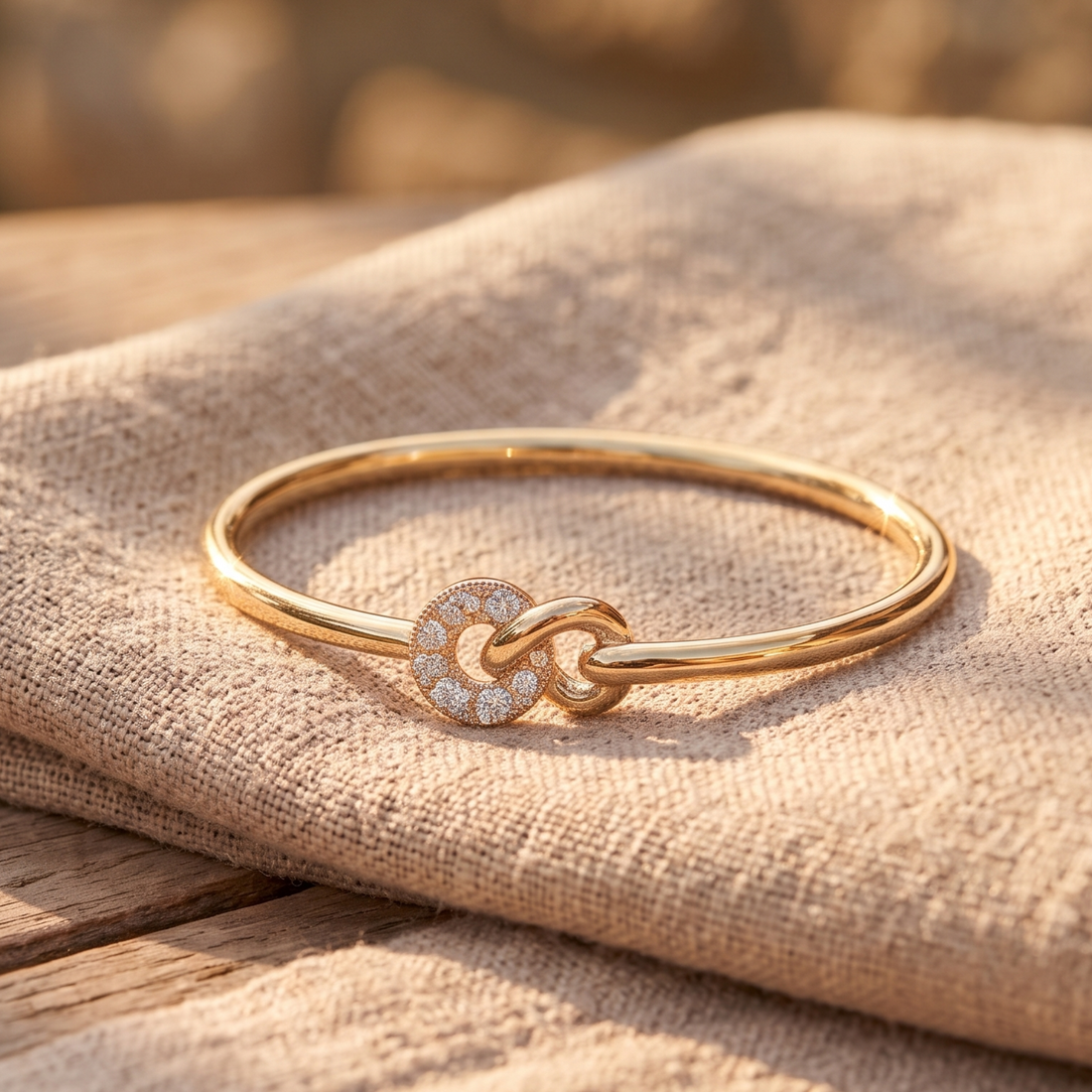 A polished gold bangle bracelet with a circular pavé-set knot charm, resting on a textured beige linen cloth over a rustic wooden surface.