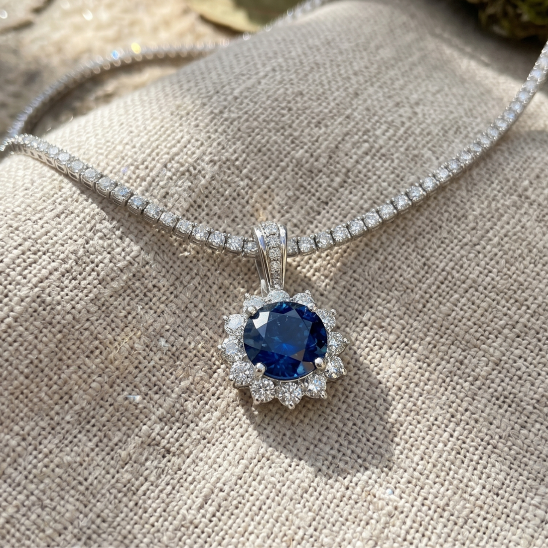 A silver-toned necklace with a round deep blue sapphire pendant, surrounded by a halo of small clear stones, resting on a textured beige fabric background.