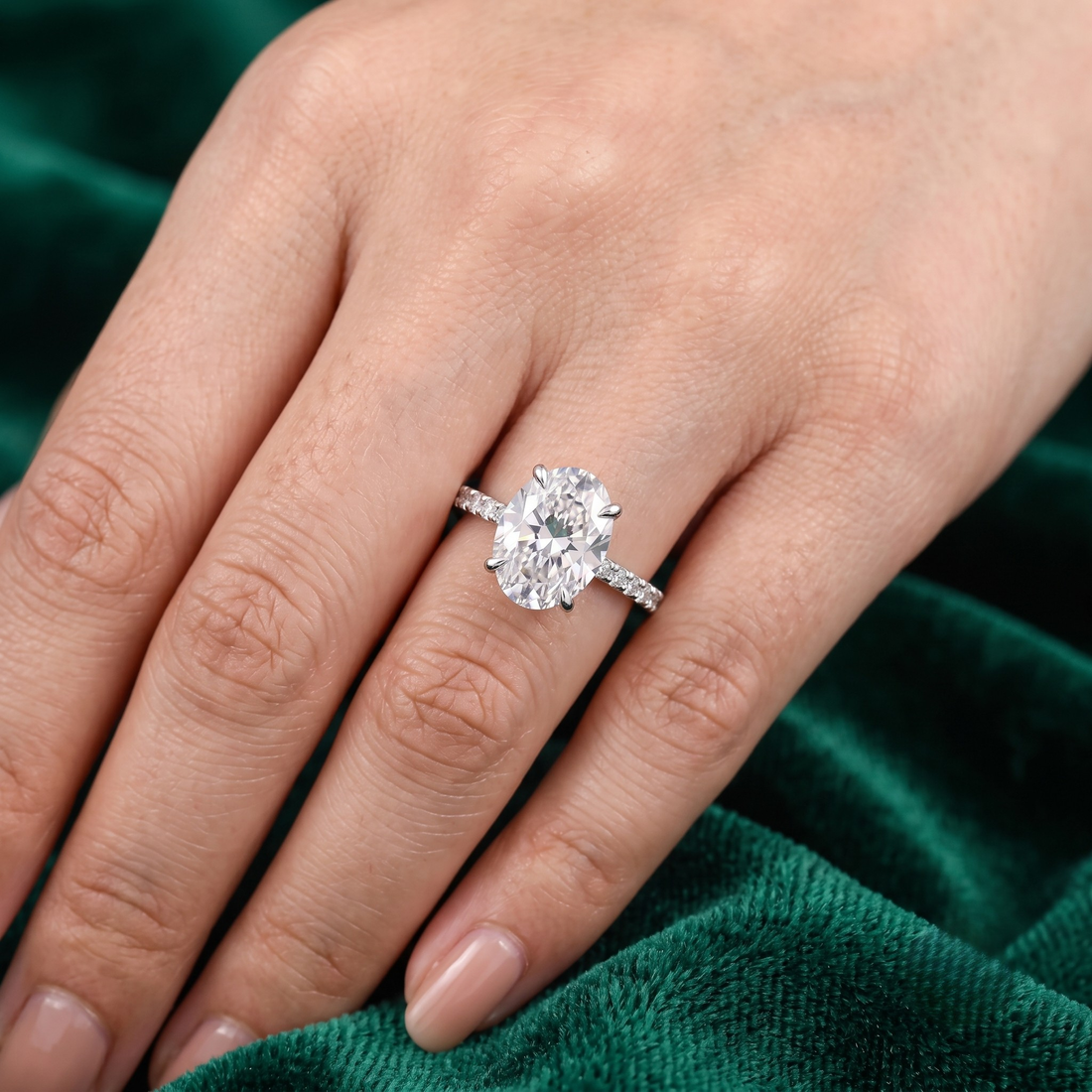 A close-up of a hand wearing a silver-toned ring with a large oval-cut clear gemstone, set against a dark green velvet fabric background.