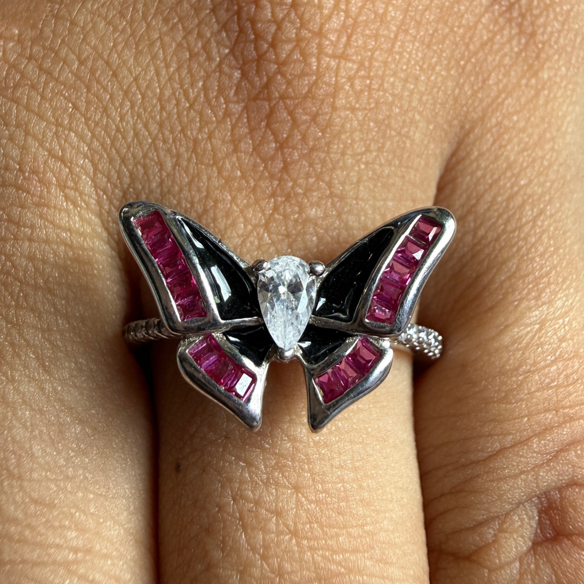 A butterfly-shaped silver ring with black enamel and bright pink gemstones, worn on a finger.
