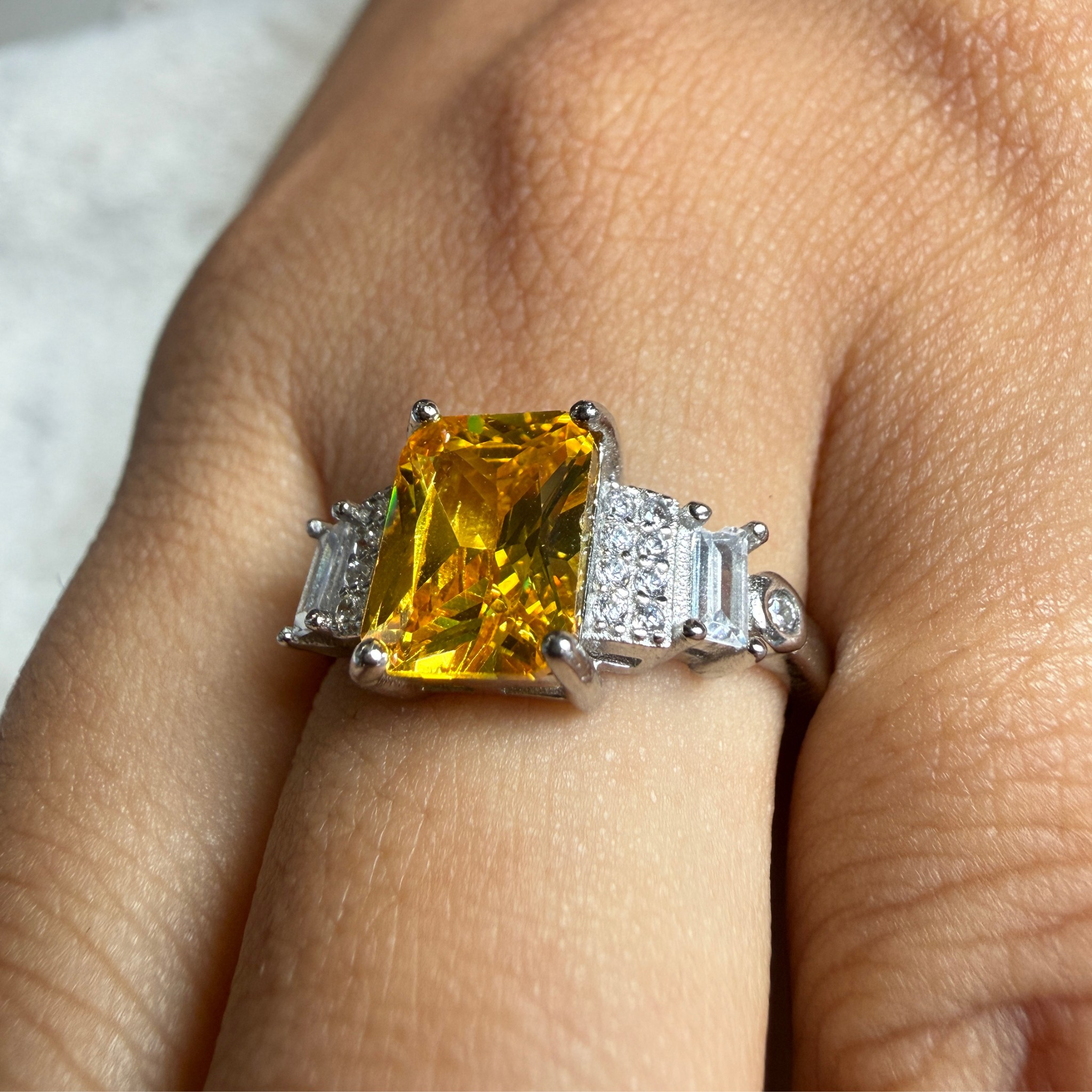 A close-up of a ring worn on a finger, featuring a large rectangular yellow gemstone in a silver-toned setting with baguette side stones.