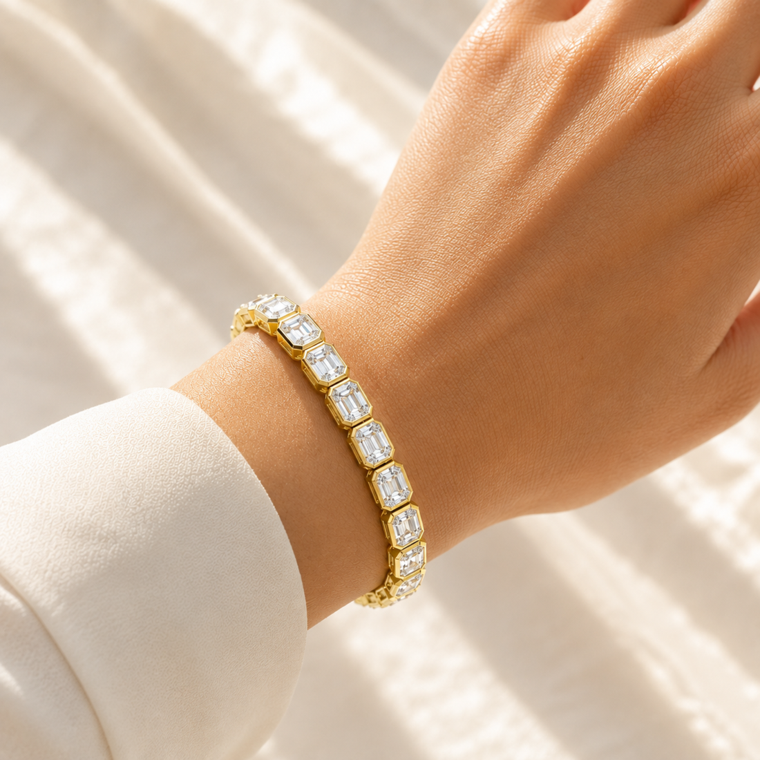A gold tennis bracelet with Emerald-cut clear stones worn on a person's wrist, shown against a soft white draped fabric background.