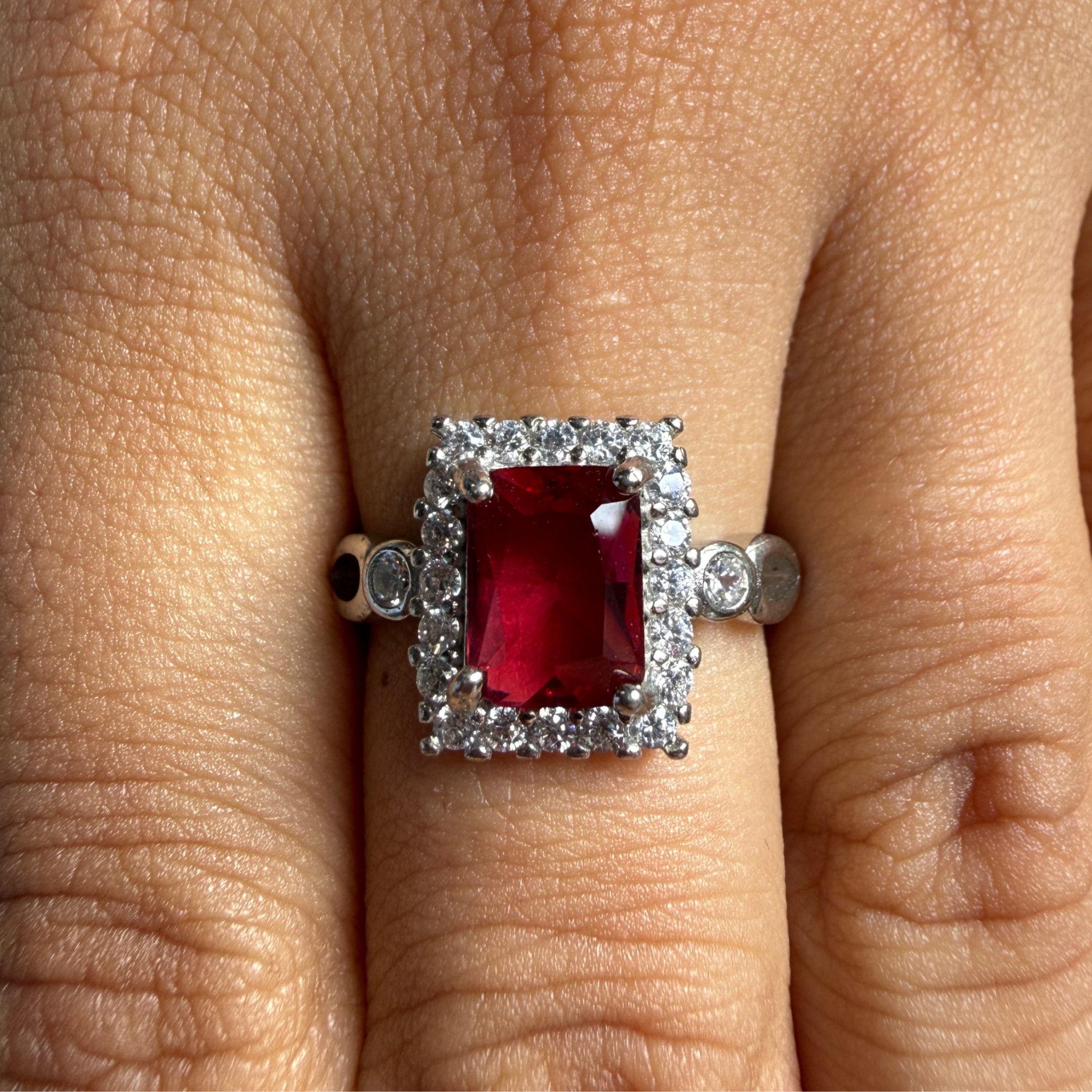 A silver ring featuring a rectangular emerald-cut red gemstone surrounded by a double halo of small clear stones, shown worn on a finger.