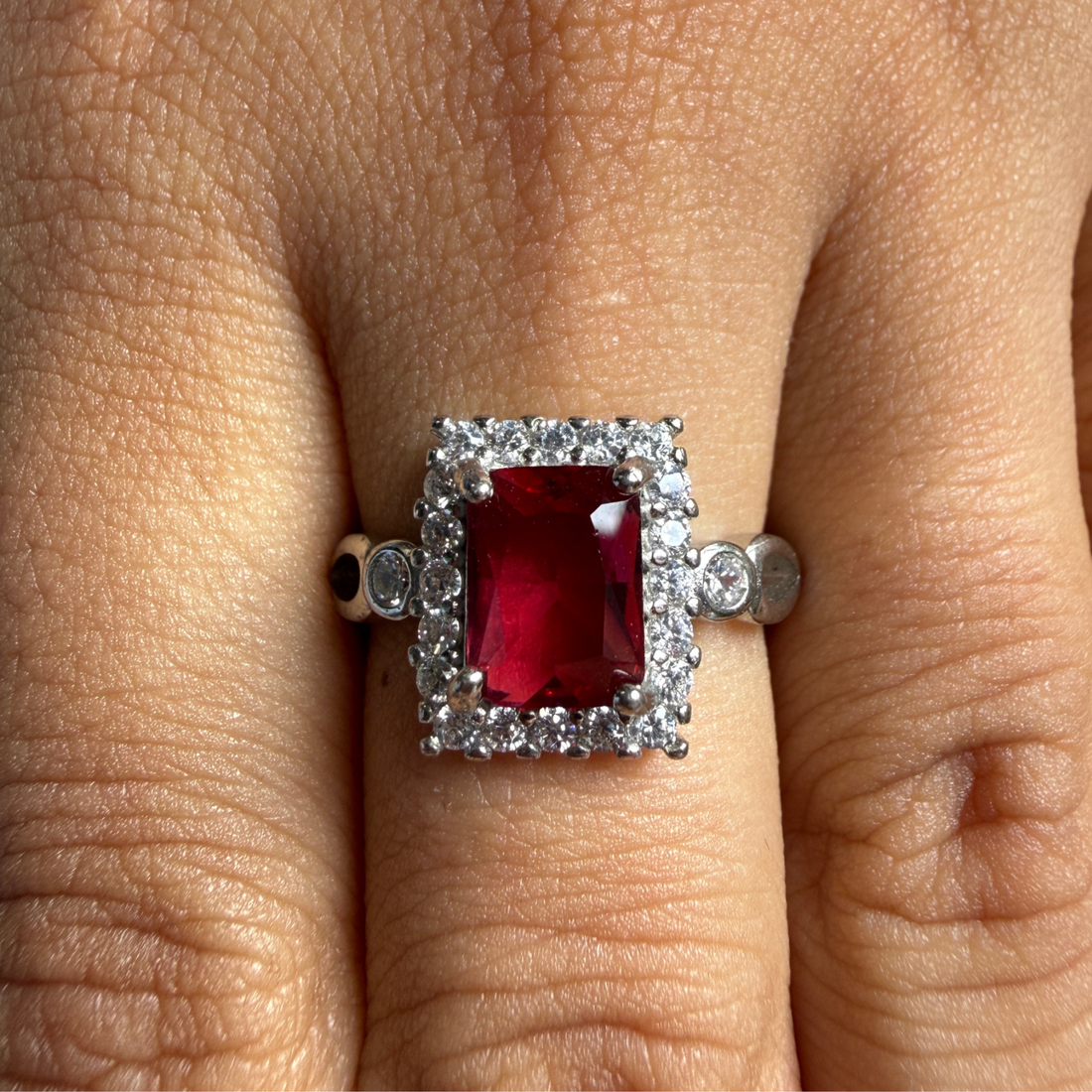 A silver ring featuring a rectangular emerald-cut red gemstone surrounded by a double halo of small clear stones, shown worn on a finger.