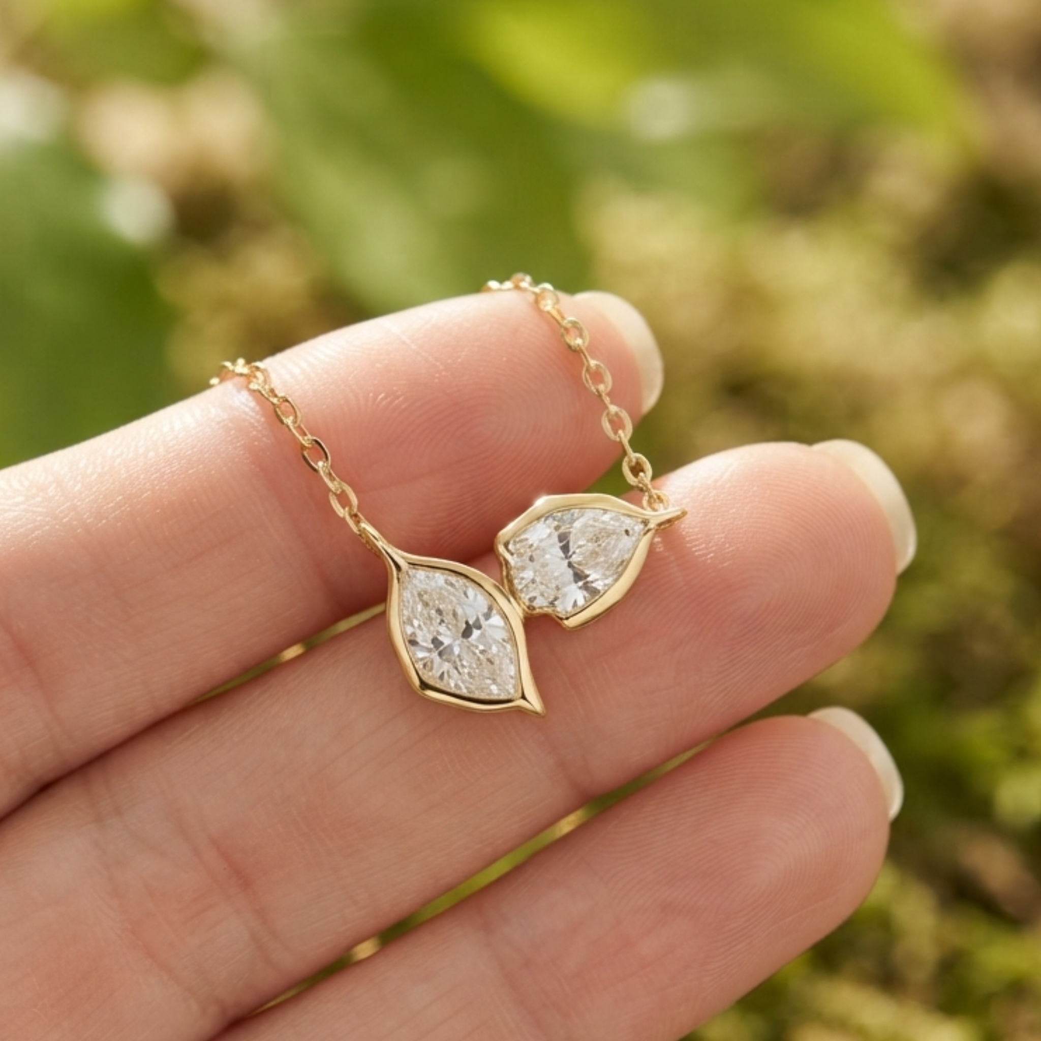 A pair of gold leaf-shaped pendant earrings with clear marquise-cut stones, held on a person's fingers against a blurred green outdoor background.