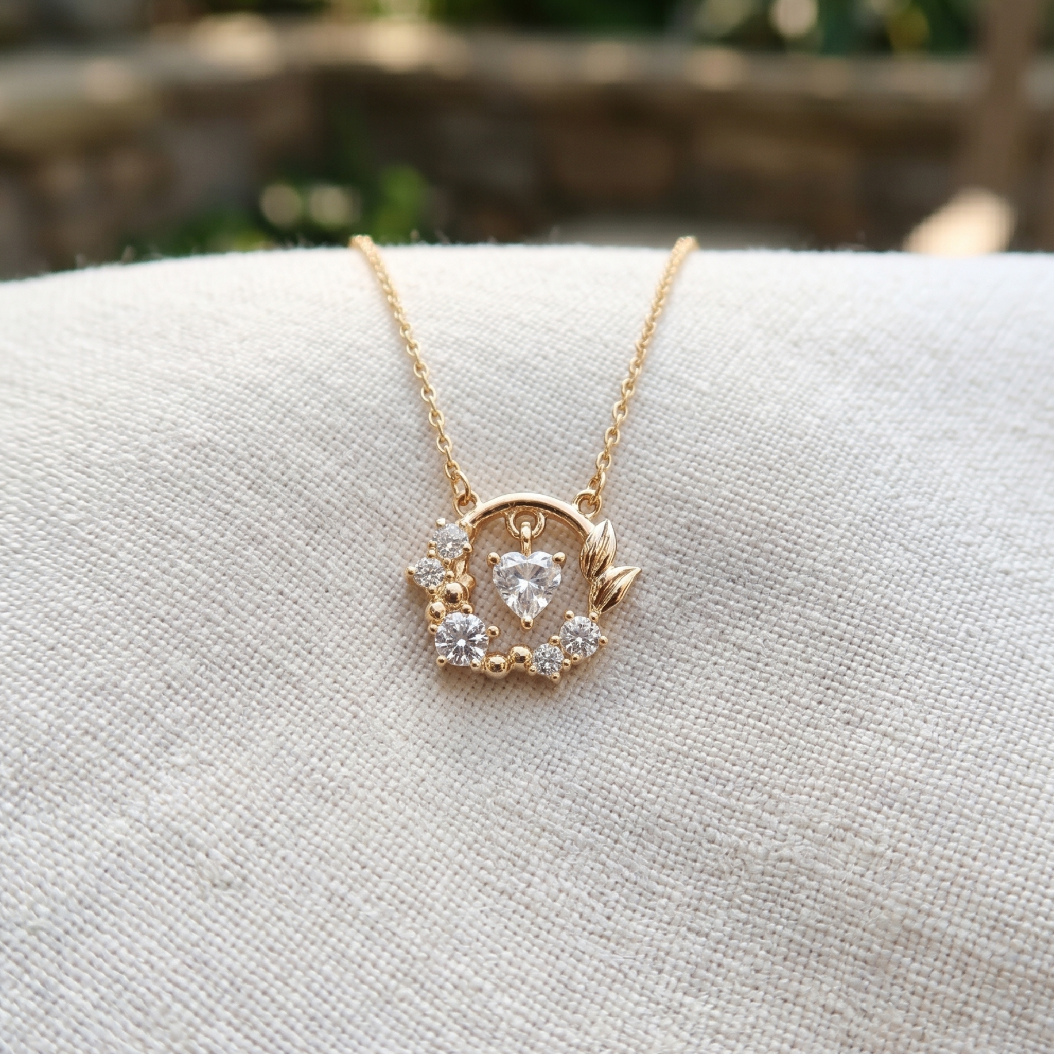 A gold heart-shaped pendant necklace with a clear heart-shaped gemstone and small clear accent stones, resting on a textured off-white fabric surface.