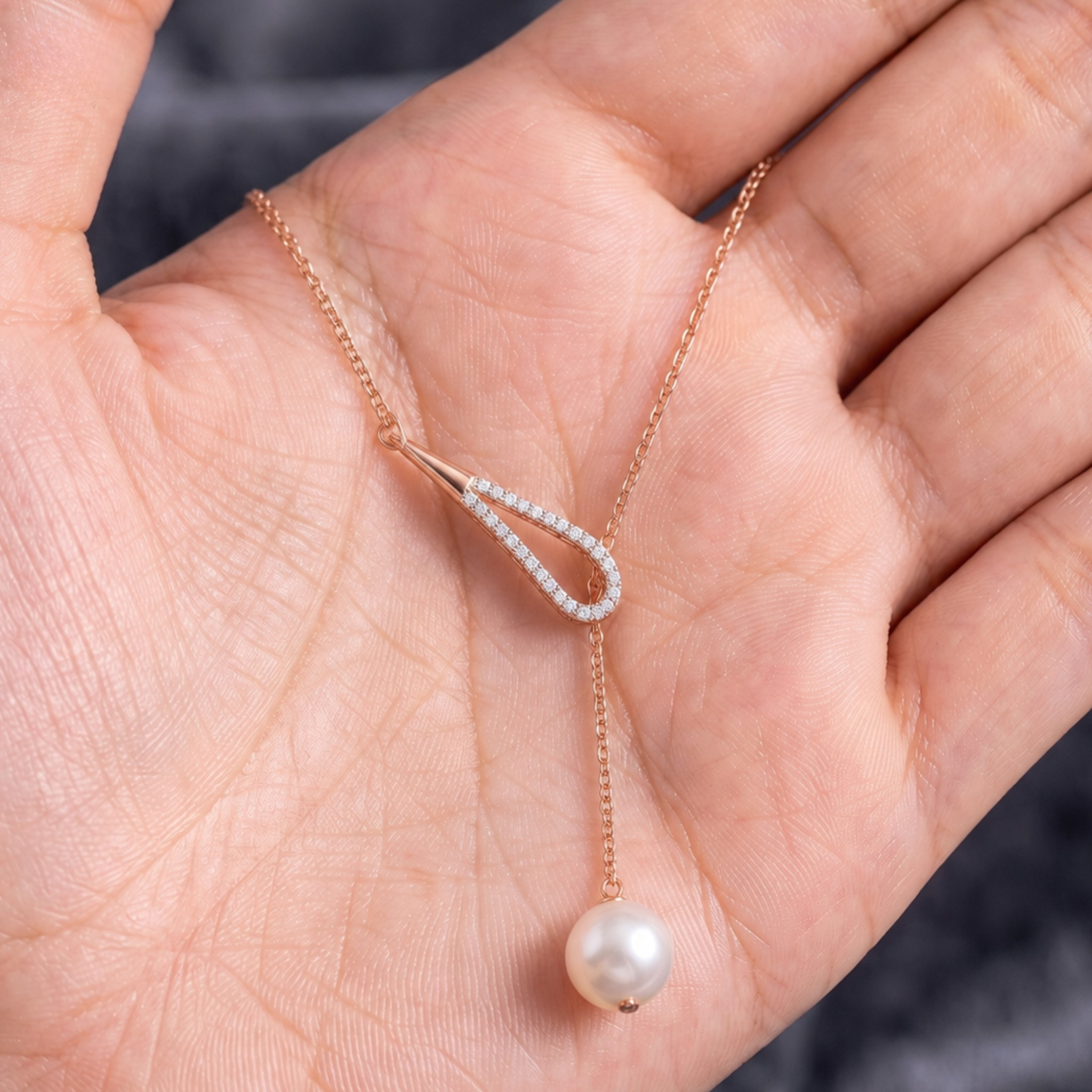 A delicate rose gold Y-necklace with a teardrop-shaped pendant set with small clear stones and a dangling white pearl, displayed on an open hand.