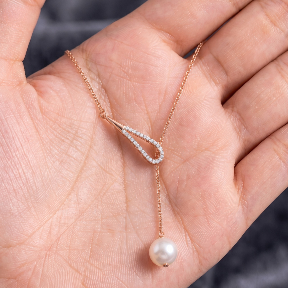 A delicate rose gold Y-necklace with a teardrop-shaped pendant set with small clear stones and a dangling white pearl, displayed on an open hand.