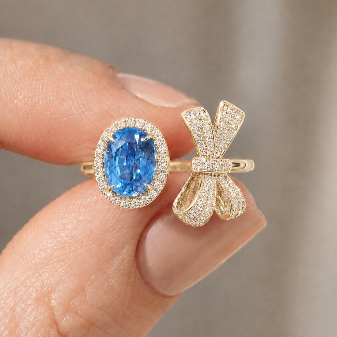 A close-up of a hand holding a yellow gold ring with an oval-cut blue sapphire gemstone surrounded by a halo of small clear stones, next to a matching bow-shaped gold ring set with clear stones.