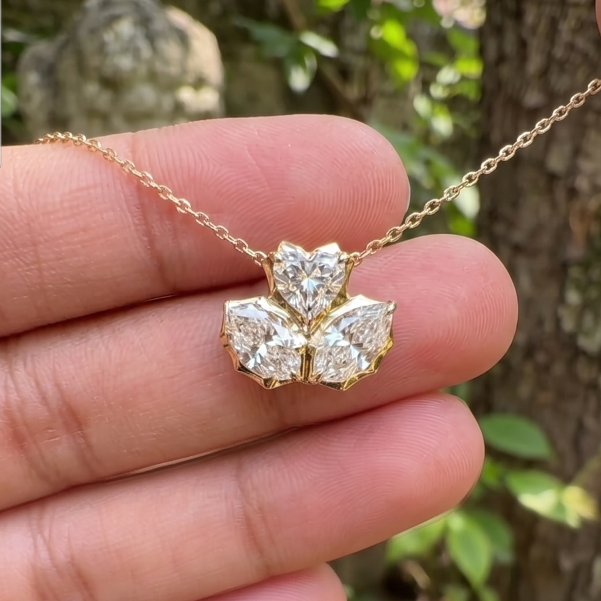 A gold necklace with a three-stone diamond pendant held in a person's hand, with a blurred outdoor background of green leaves and tree bark.