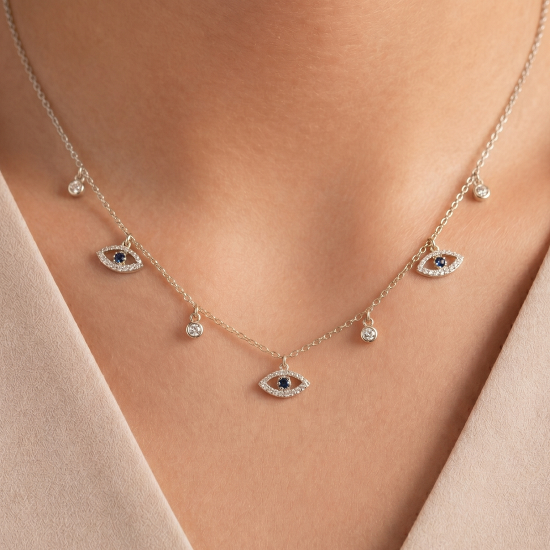 A delicate gold chain necklace with three evil eye charms and small teardrop beads, worn on a model's neck. The evil eye charms feature dark blue centers surrounded by clear stones, and the necklace is styled with a beige top.