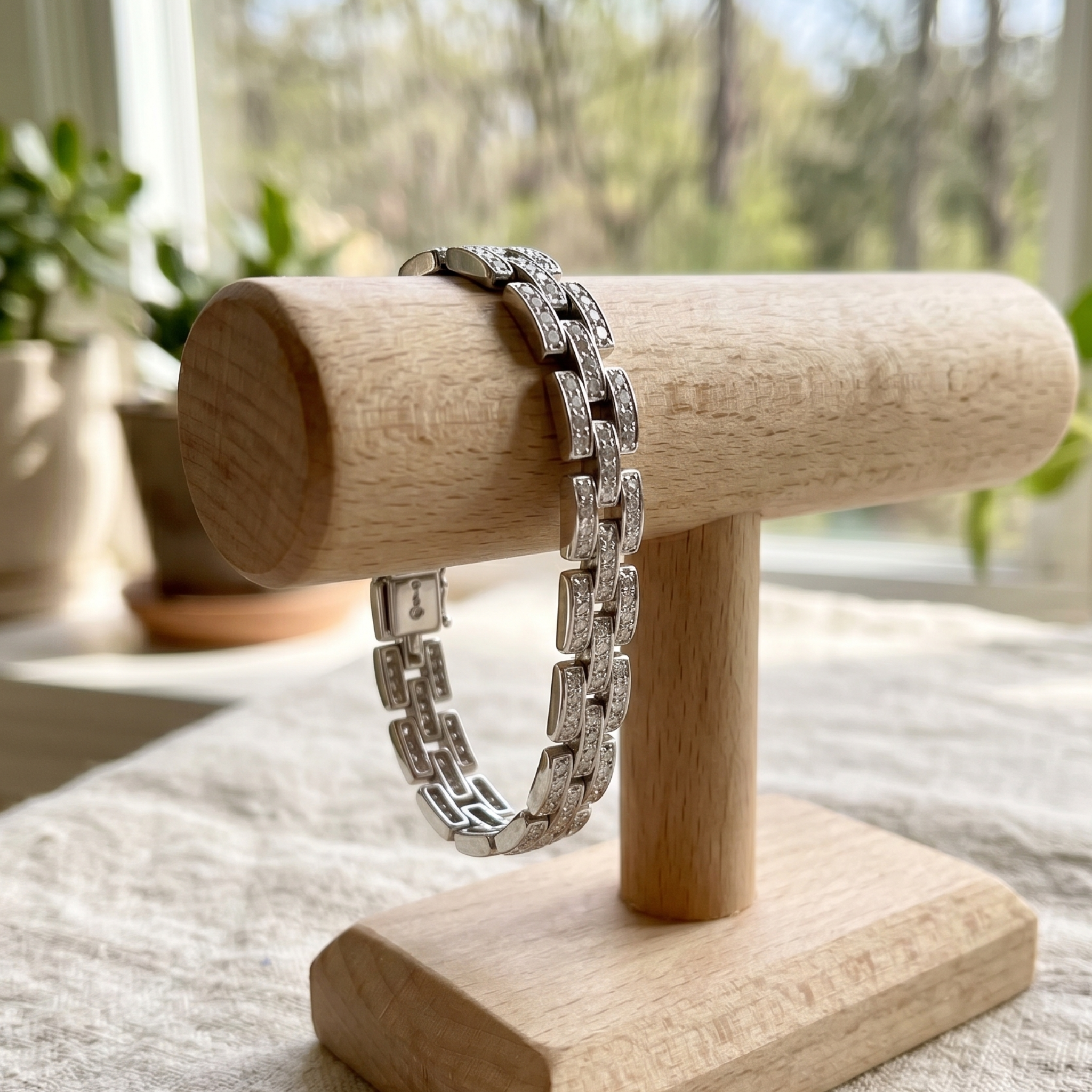 A silver-toned link bracelet displayed on a light wood jewelry stand, with a softly blurred indoor background featuring plants and a window.