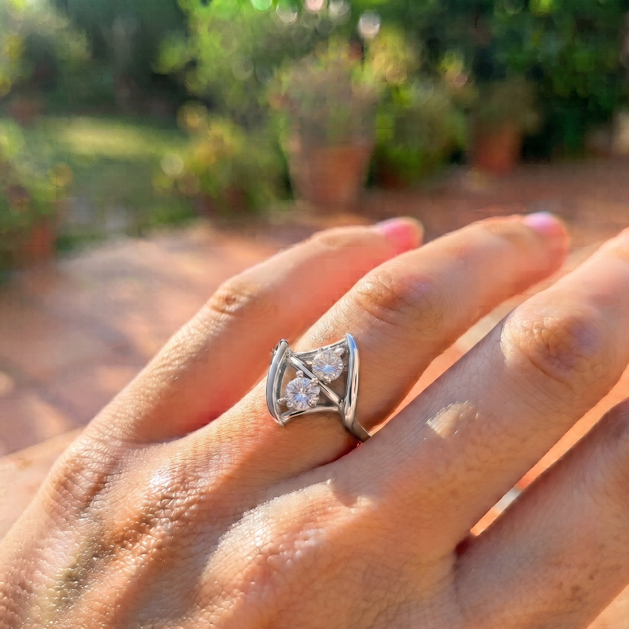 A silver ring with a geometric triangular design and clear gemstones, worn on a person's hand against a blurred outdoor garden background.