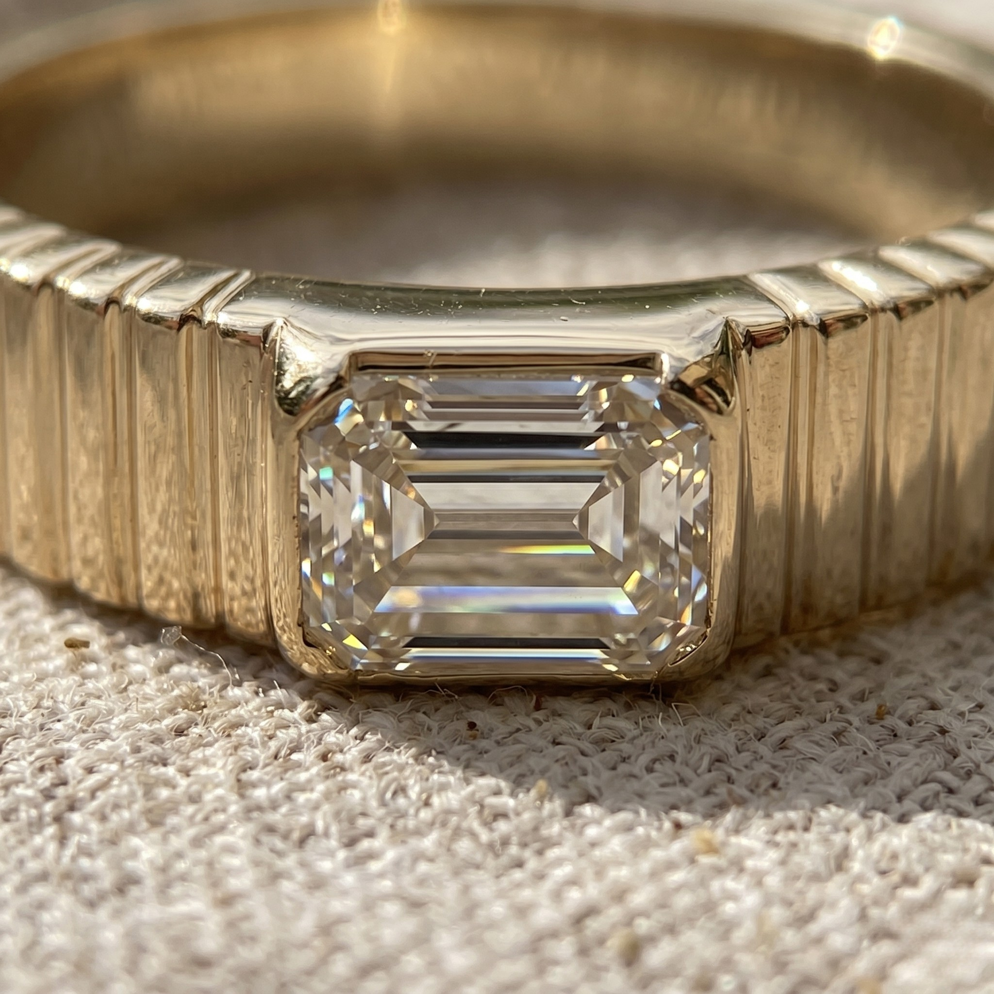 A close-up of a yellow gold ring featuring an emerald-cut clear gemstone in a prong setting, resting on a textured beige fabric surface.
