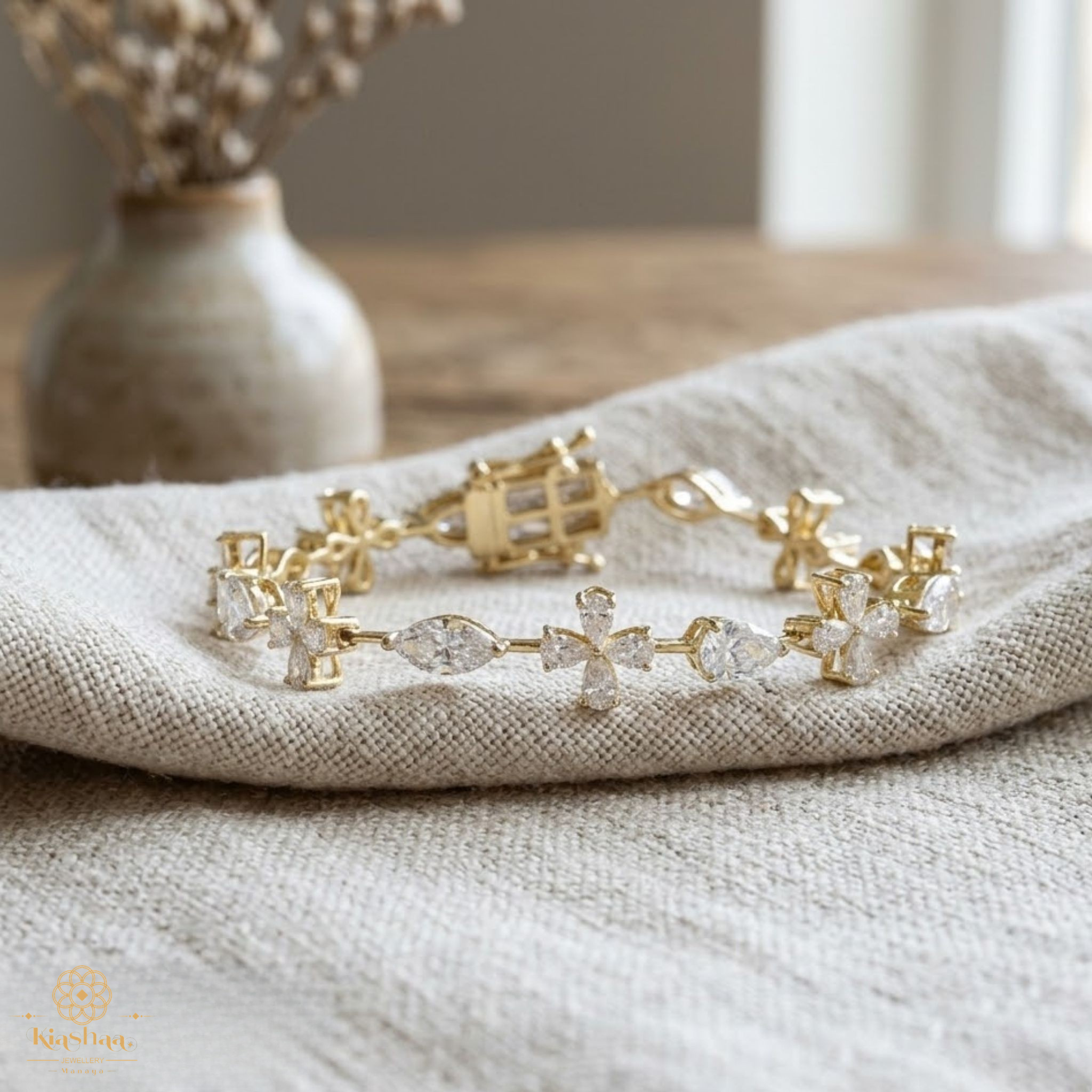 A gold bracelet with clear gemstones, including a central cross motif, resting on a textured beige linen cloth with a blurred vase in the background.