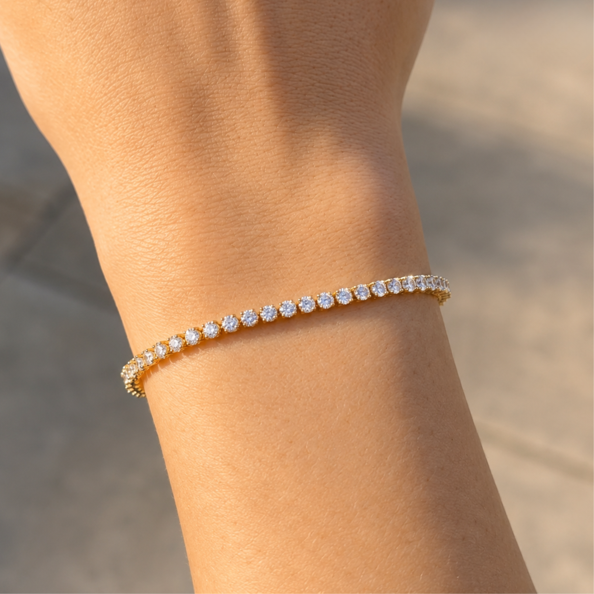 A delicate gold tennis bracelet with small clear stones, shown worn on a person's wrist against a softly blurred outdoor background.