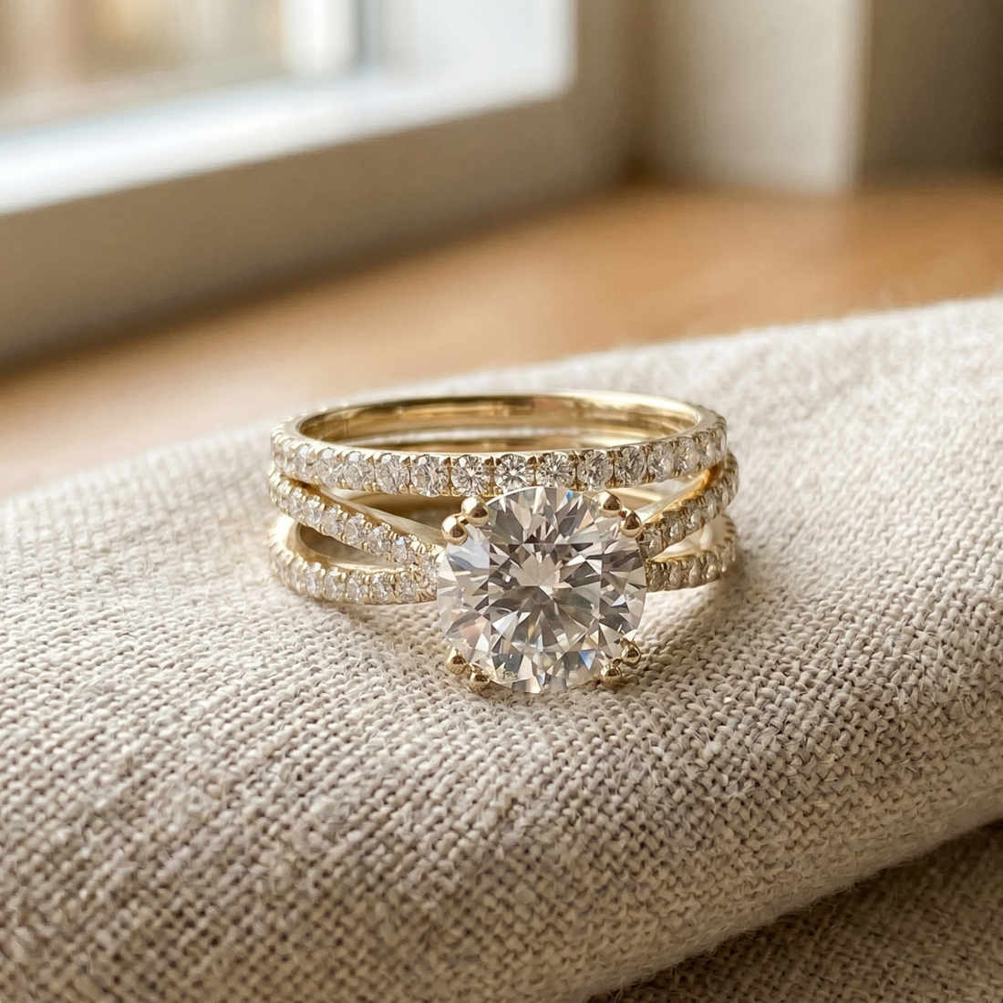 A gold ring set with a large round-cut center stone and smaller pavé-set stones, displayed on a textured beige fabric surface near a softly blurred window.
