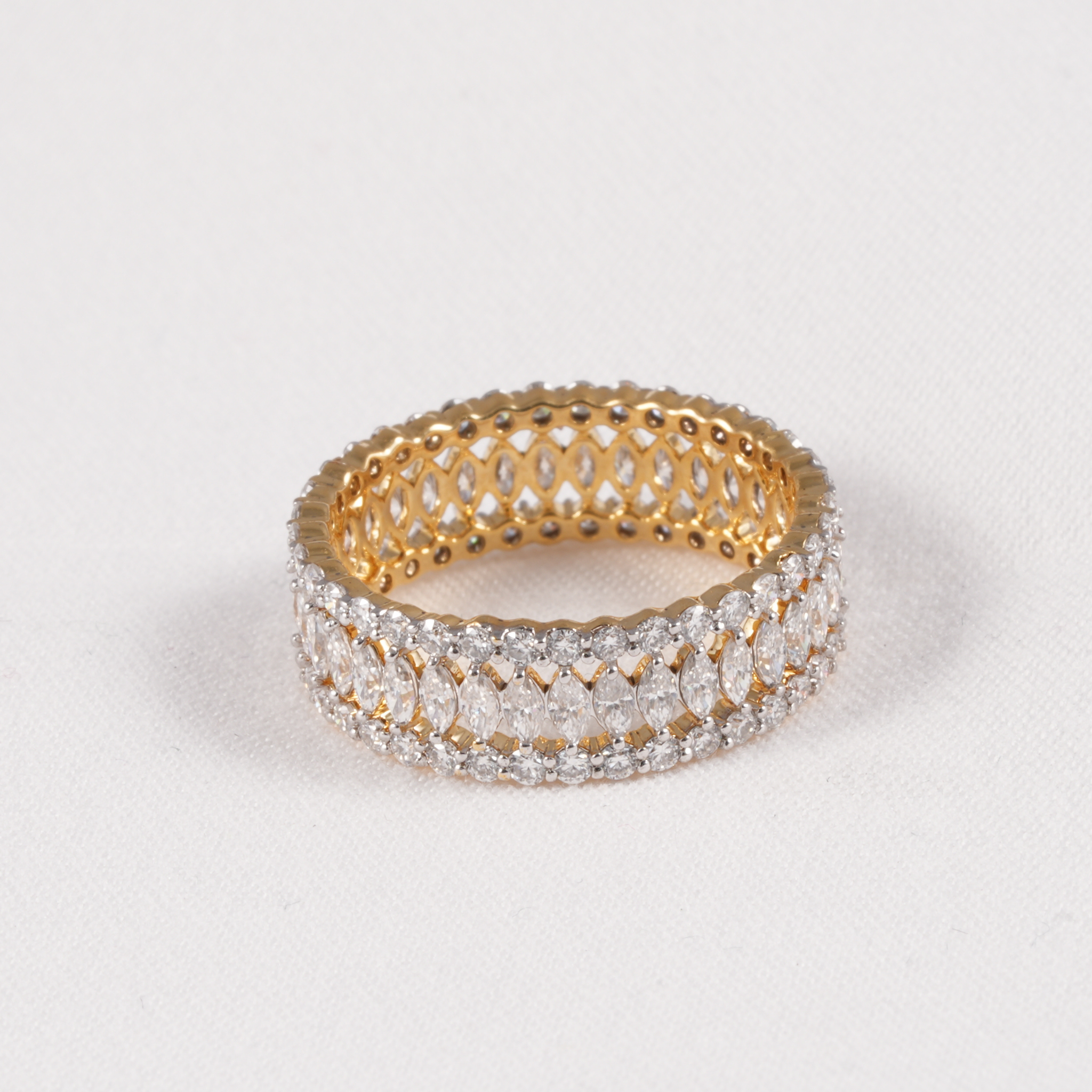 A gold eternity ring featuring three rows of clear pavé-set round and marquise-cut gemstones, photographed on a light gray textured background.