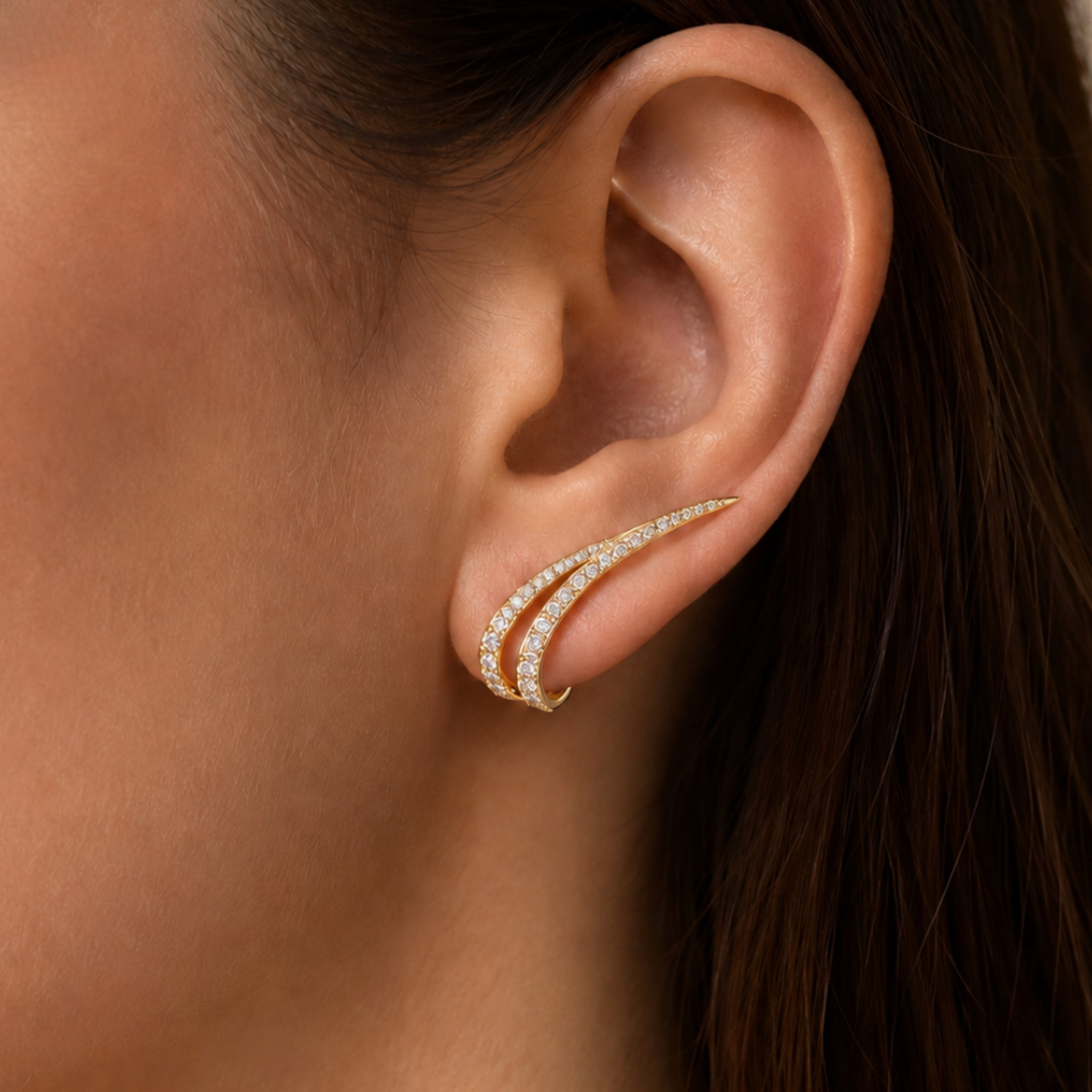 A close-up of a model's ear wearing a delicate gold ear cuff adorned with small clear pavé stones. The earrings are shown in warm, direct lighting against the model's skin and dark brown hair.