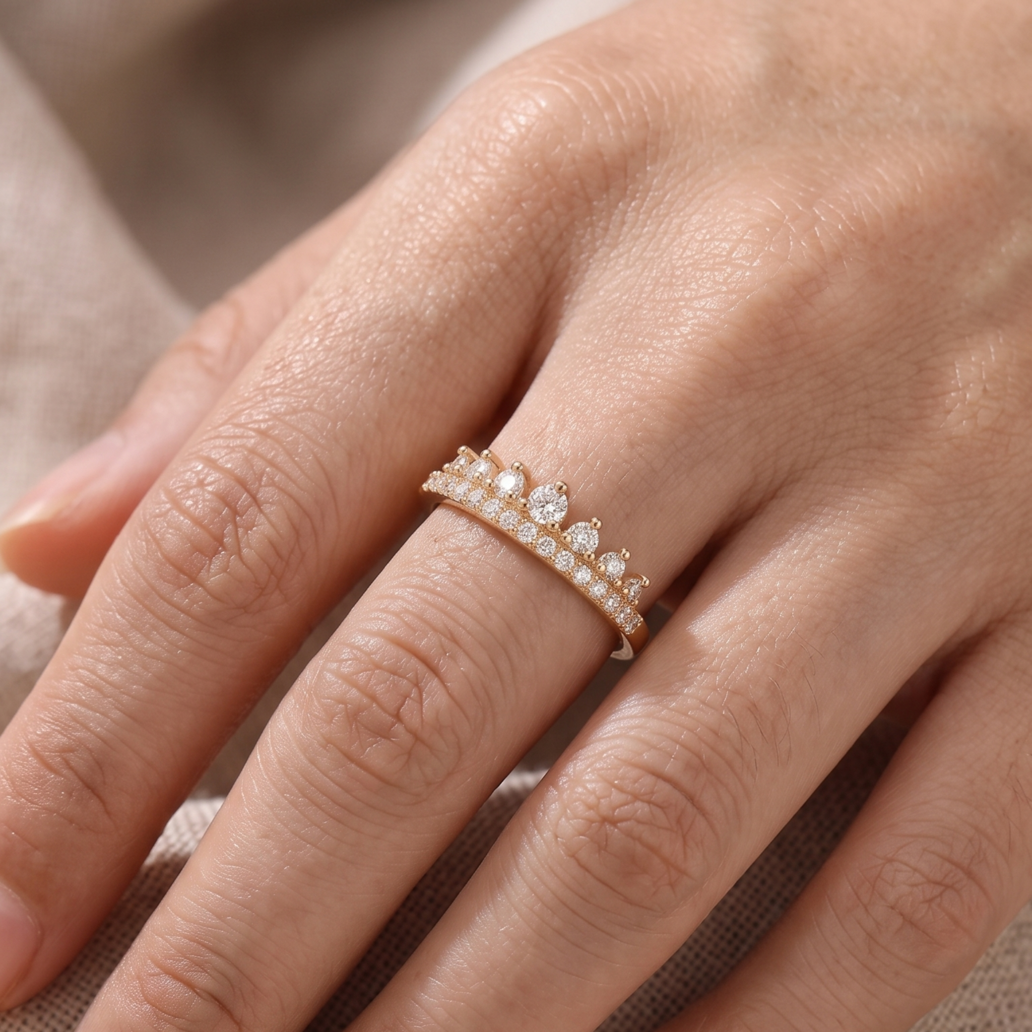 A close-up of a hand wearing a delicate gold ring adorned with small clear stones and a central round gemstone, set against a soft neutral fabric background.