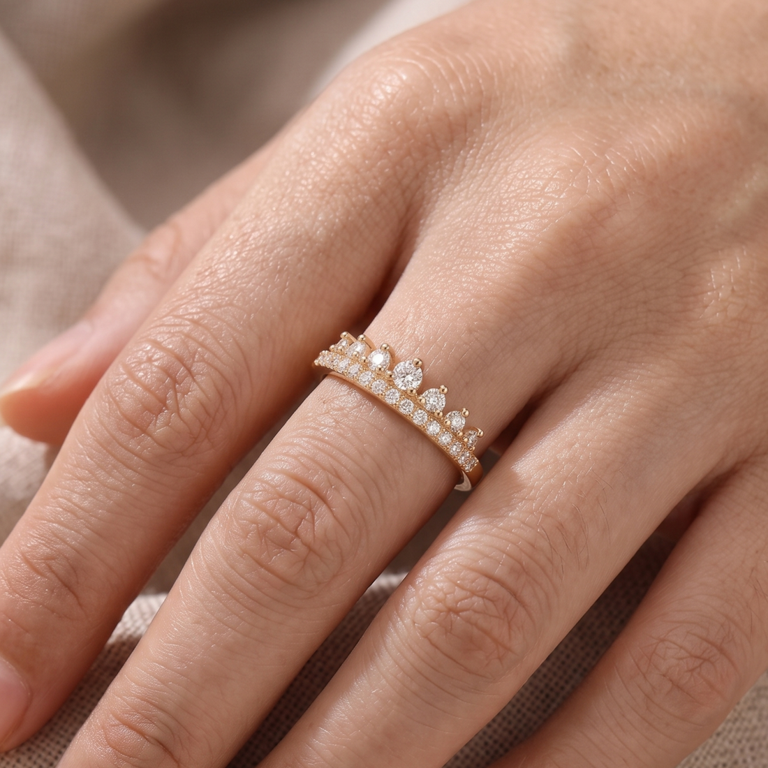 A close-up of a hand wearing a delicate gold ring adorned with small clear stones and a central round gemstone, set against a soft neutral fabric background.