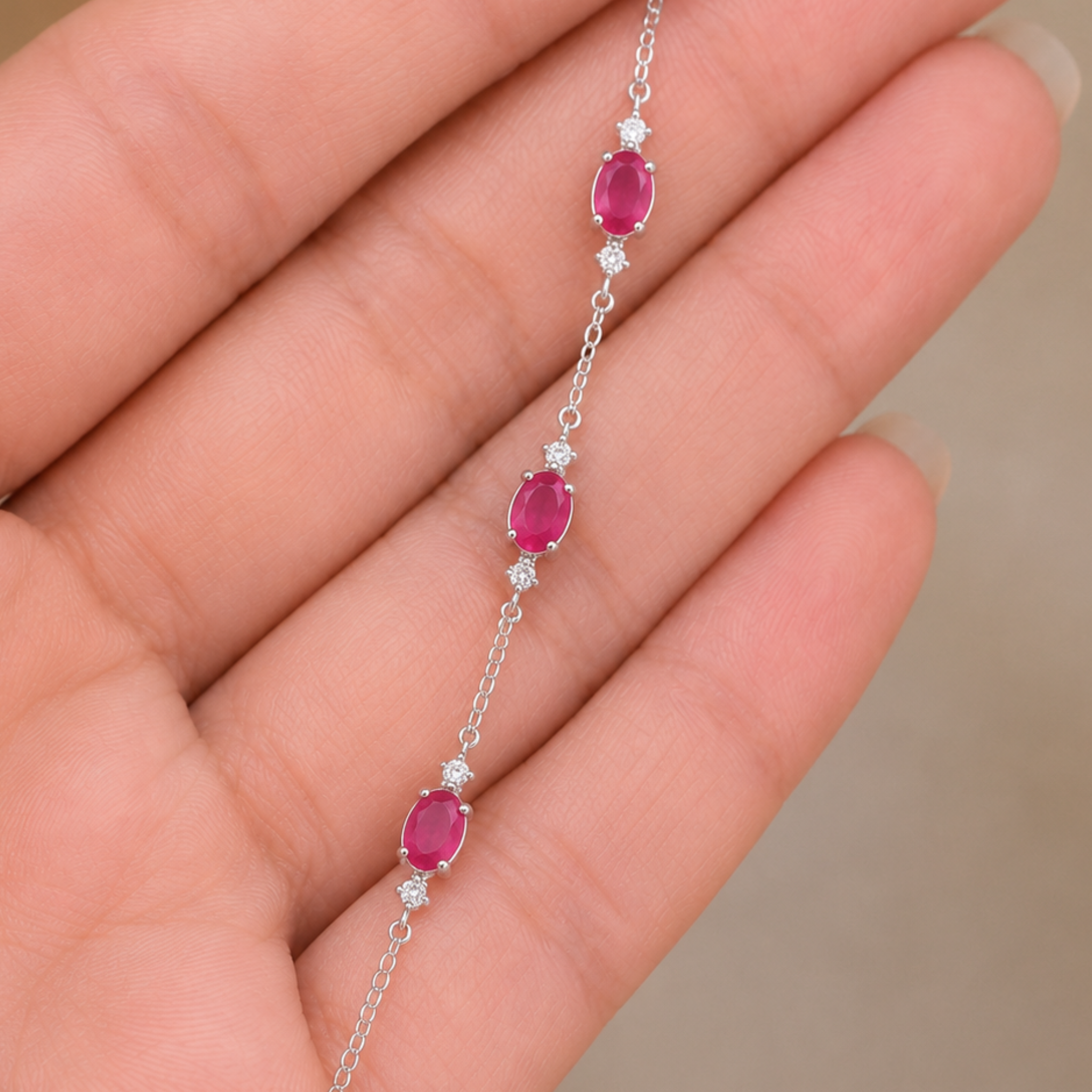 A delicate silver chain bracelet with three oval pinkish-red gemstones and small clear accent stones, shown resting on a person's hand.