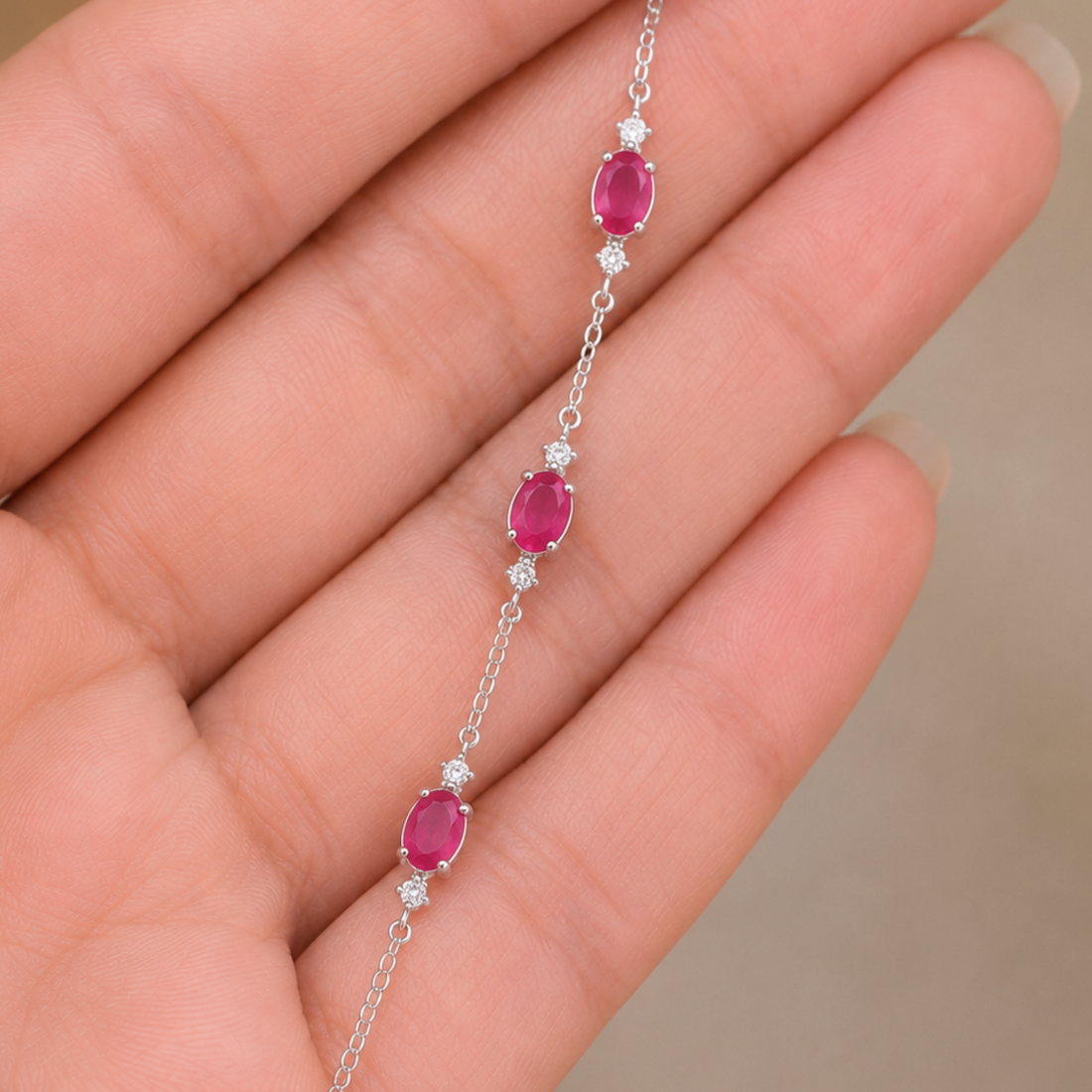 A delicate silver chain bracelet with three oval pinkish-red gemstones and small clear accent stones, shown resting on a person's hand.