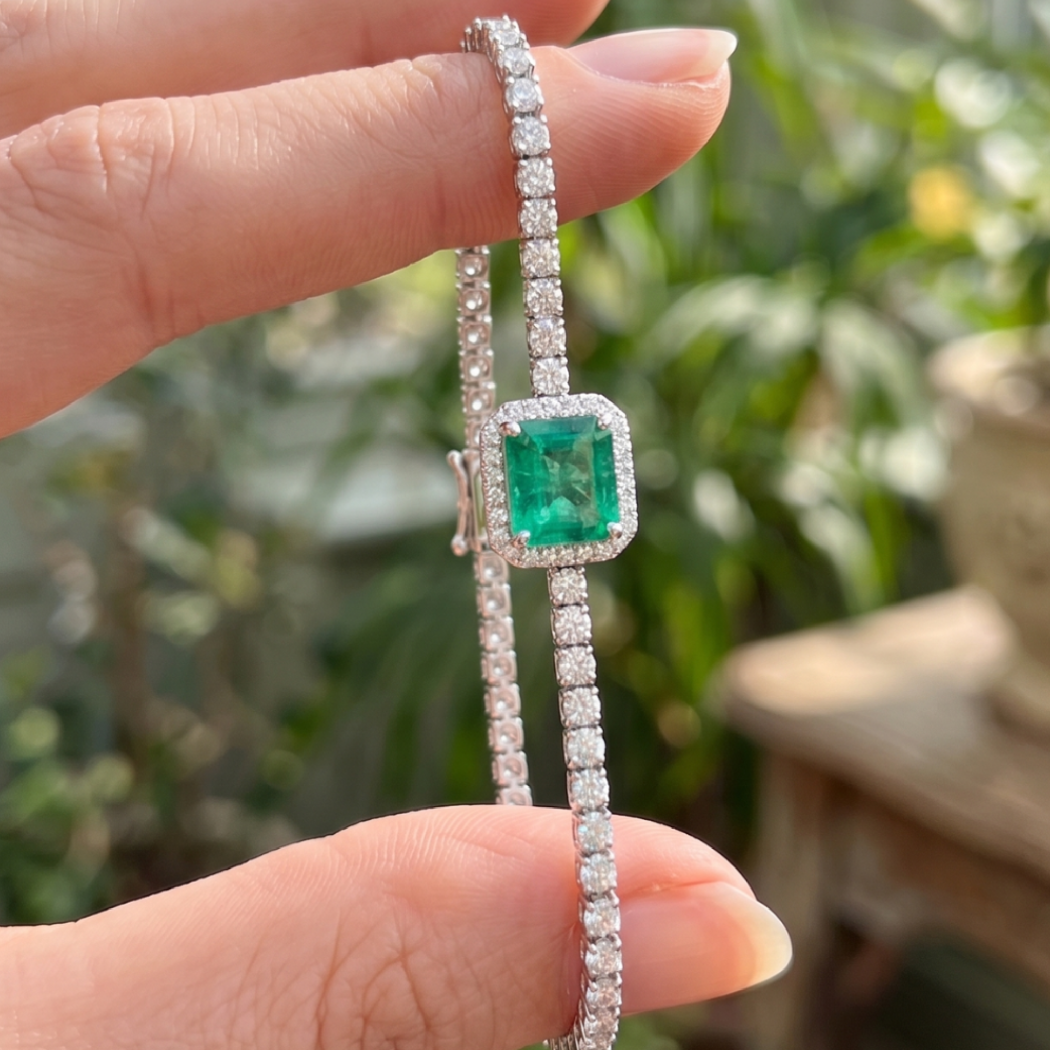 A close-up of a hand holding a silver-toned bracelet with a square-cut green emerald gemstone surrounded by small clear stones, set against a blurred outdoor background.