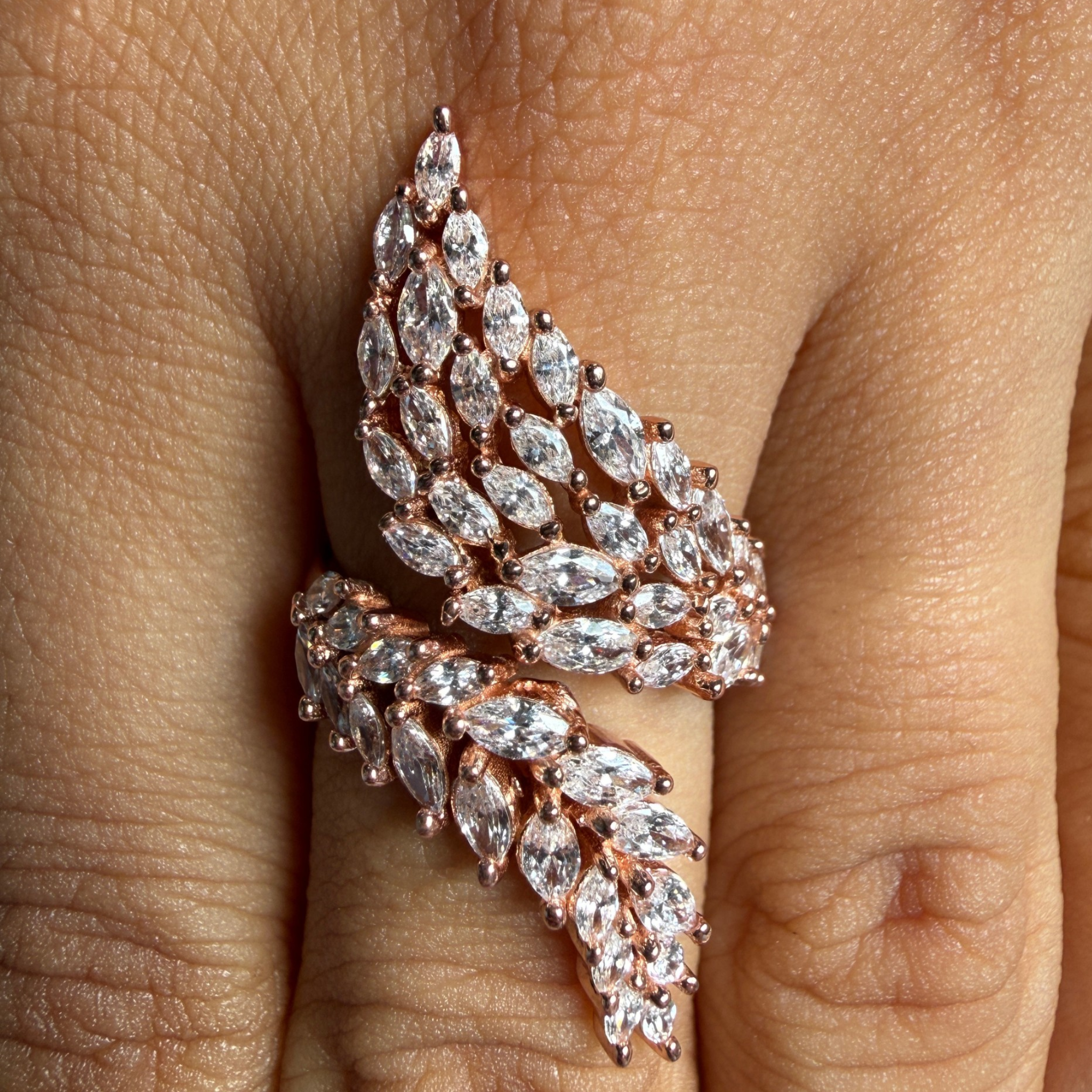 A close-up of a rose gold ring with marquise-cut clear crystals arranged in a leaf or wing-like pattern, worn on a finger.