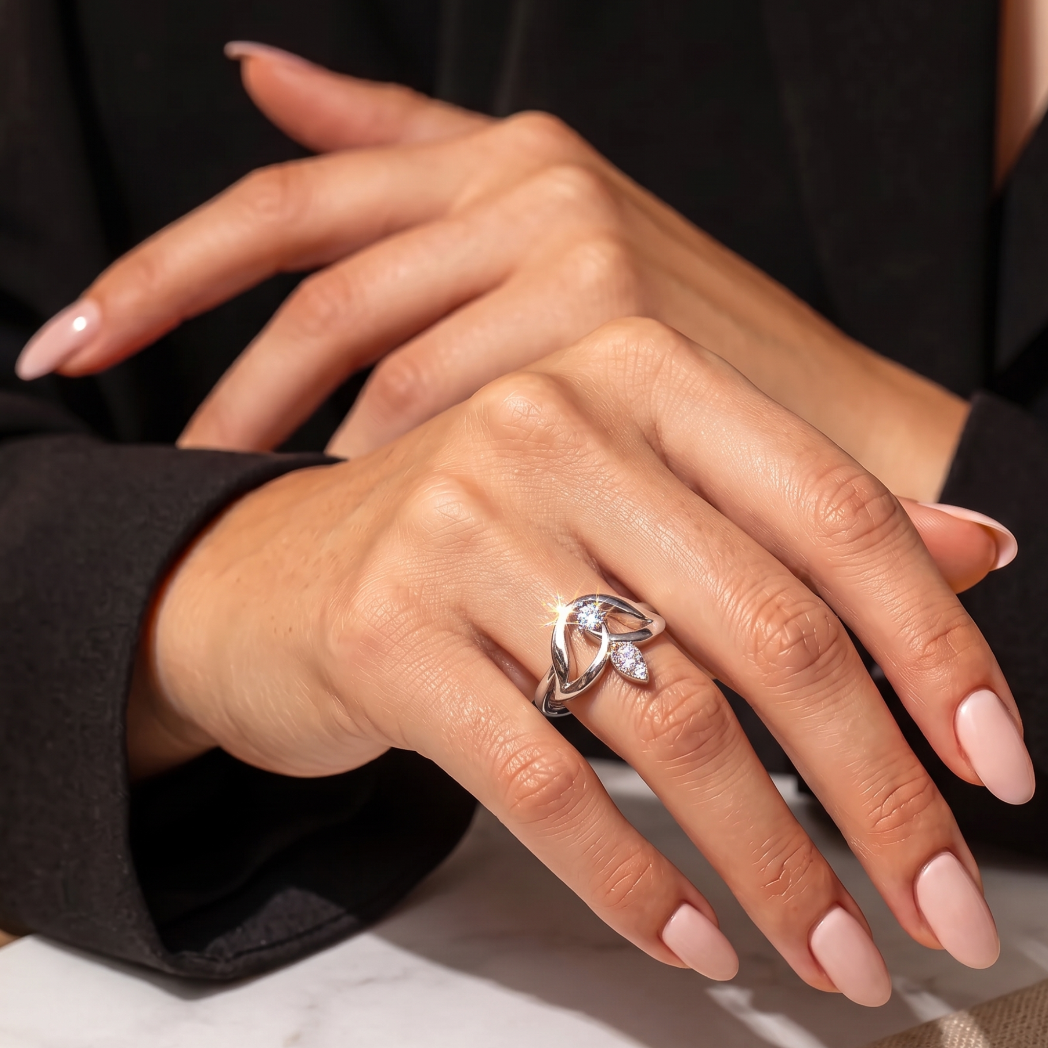 A close-up of a person's hands wearing a silver-toned ring with a small clear gemstone, styled with a black blazer and pale pink manicured nails.
