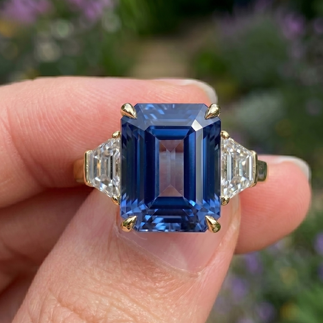 A close-up of a yellow gold ring featuring a large emerald-cut blue sapphire center stone and baguette side stones, held between fingers against a blurred garden background.