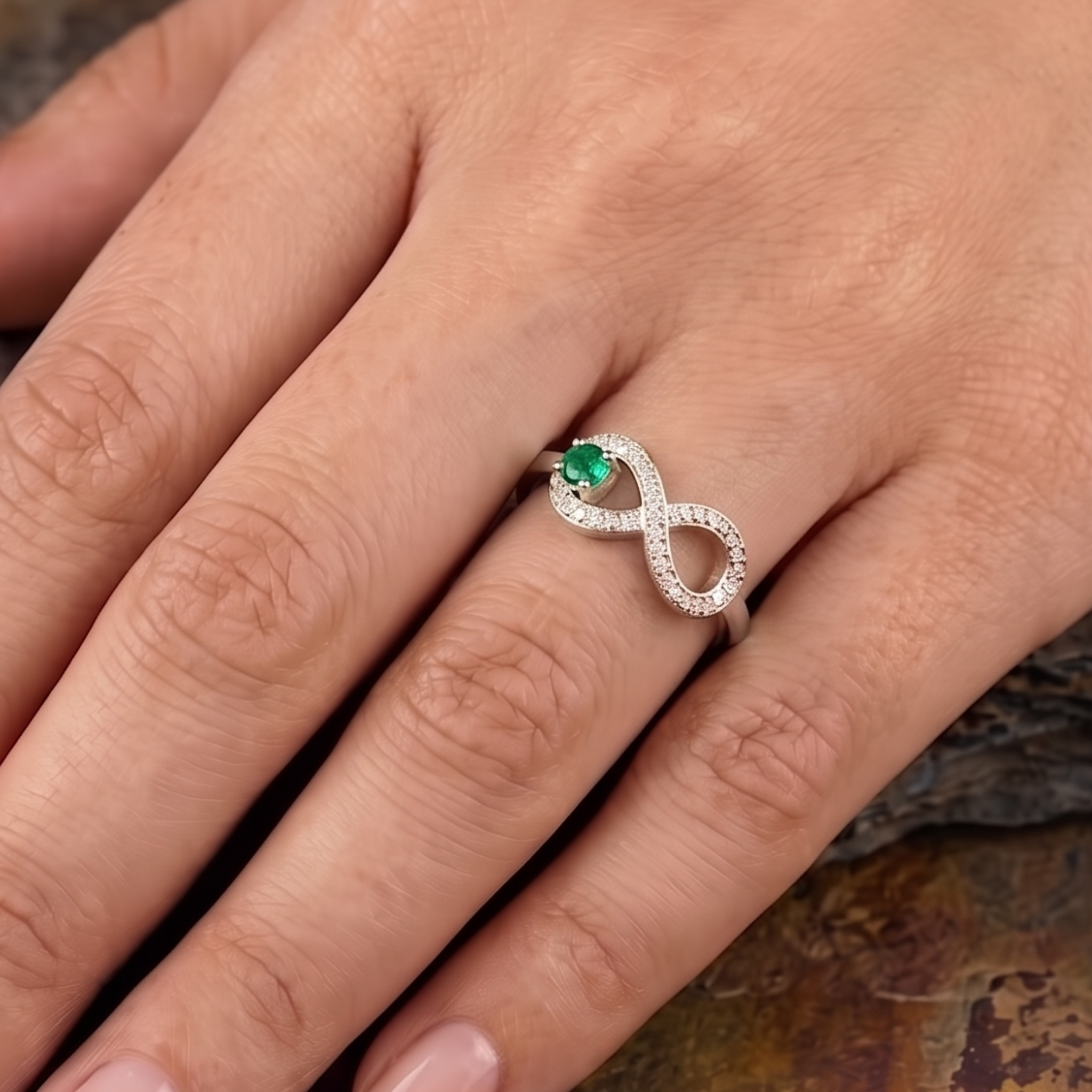 A silver infinity ring with a green emerald center stone and small clear accent stones, shown worn on a hand.
