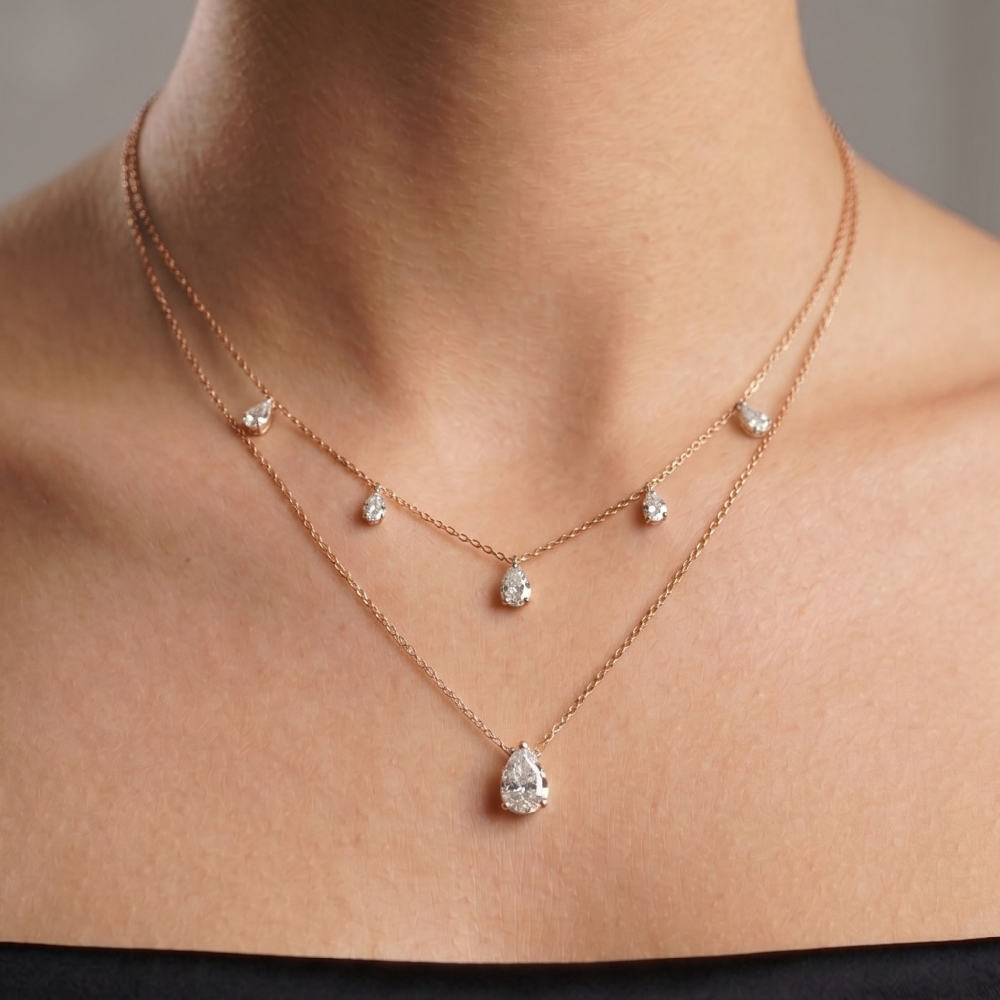 A close-up of a model's neck and upper chest wearing a delicate rose gold double-layer necklace with clear teardrop-cut gemstones.