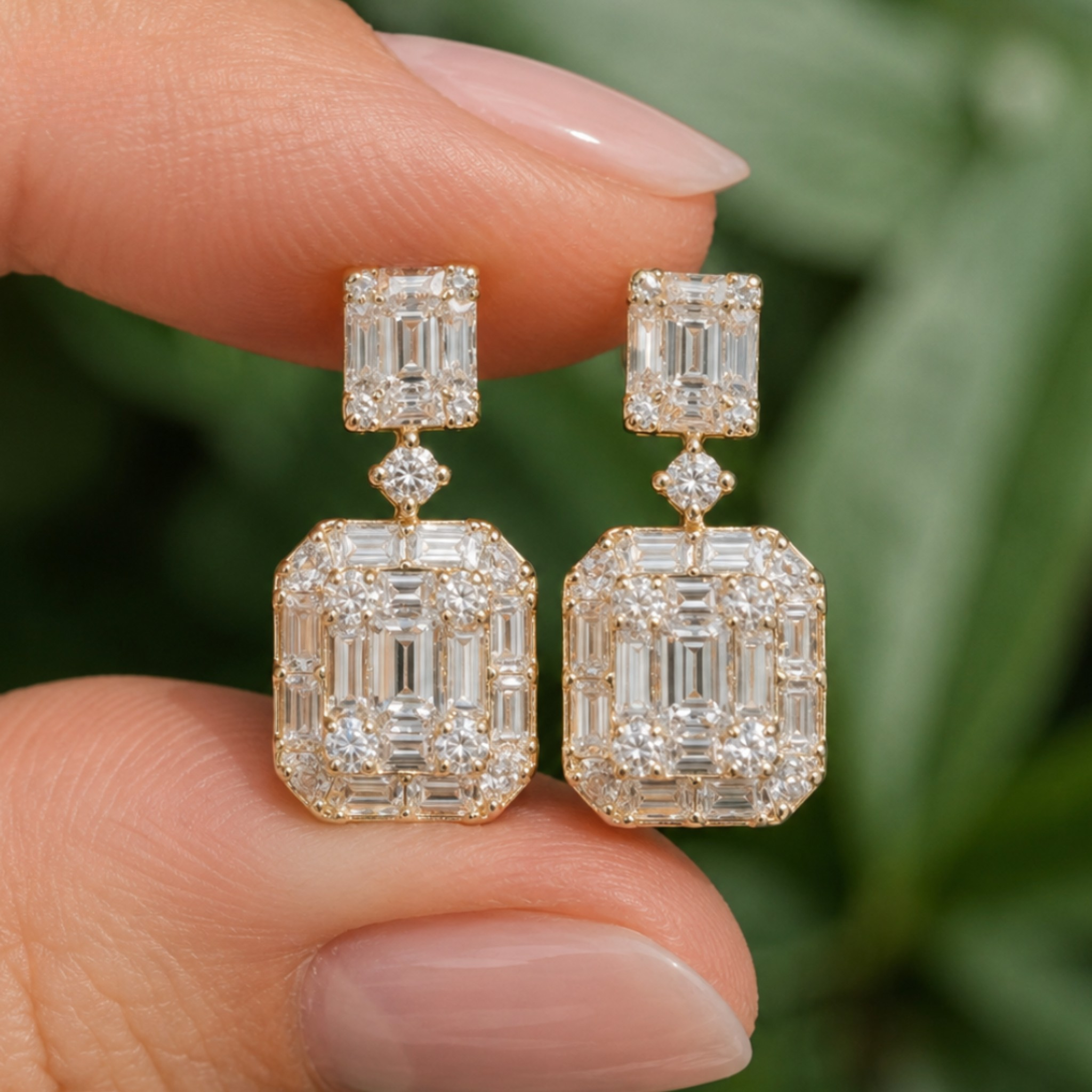 A pair of gold drop earrings with rectangular emerald-cut clear gemstones, held between two fingers against a soft green leaf background.