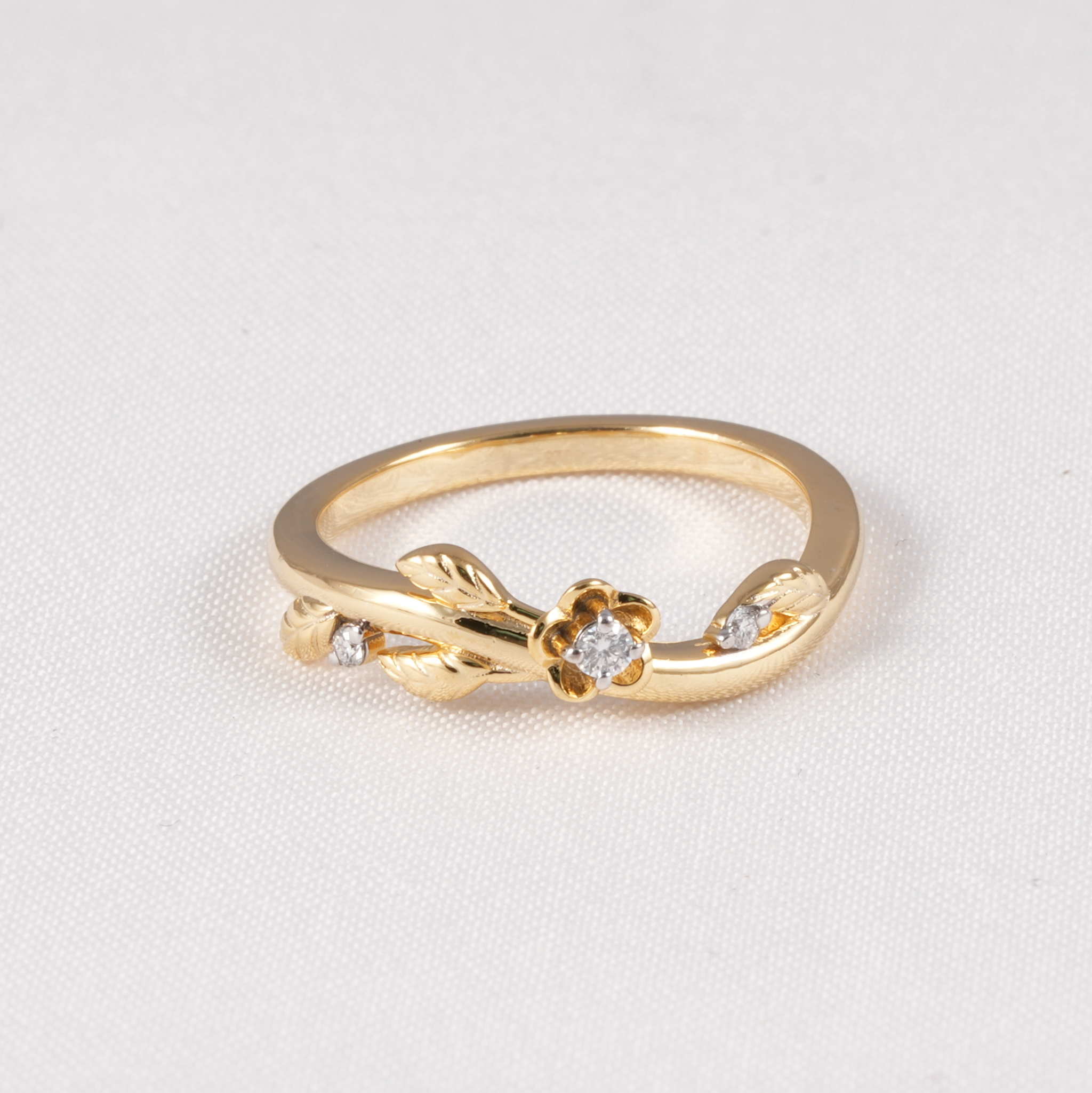 A gold ring featuring a small flower motif with a central clear gemstone and two side stones, accented by leaf-like details and small side stones, set on a textured white background.