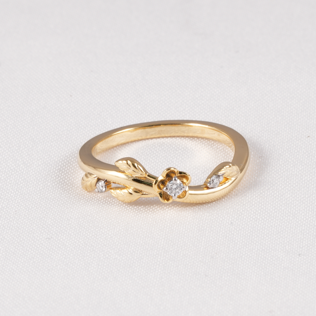 A gold ring featuring a small flower motif with a central clear gemstone and two side stones, accented by leaf-like details and small side stones, set on a textured white background.