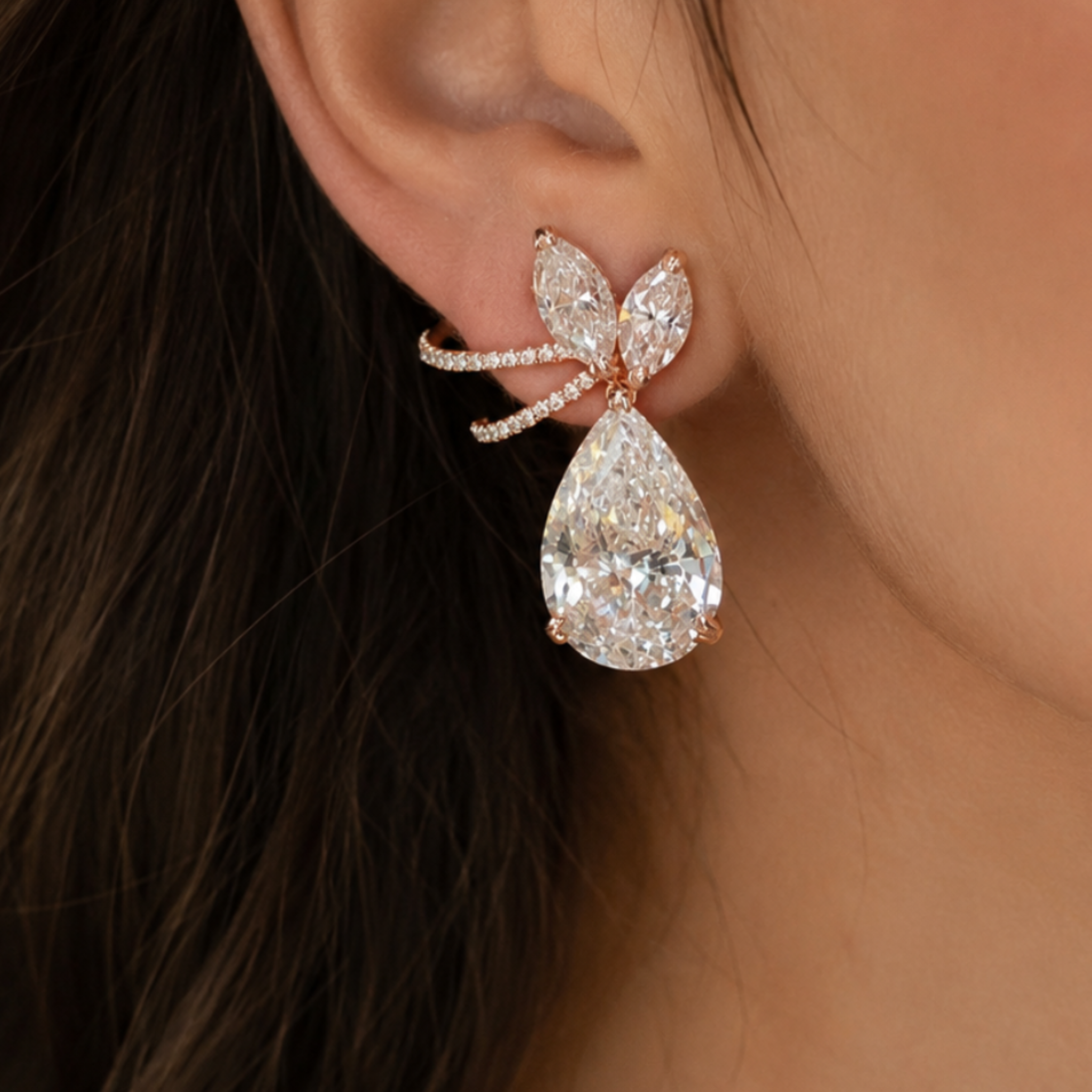 A close-up of a person's ear wearing a rose gold bow-shaped stud earring adorned with clear crystals and a teardrop-shaped crystal drop.