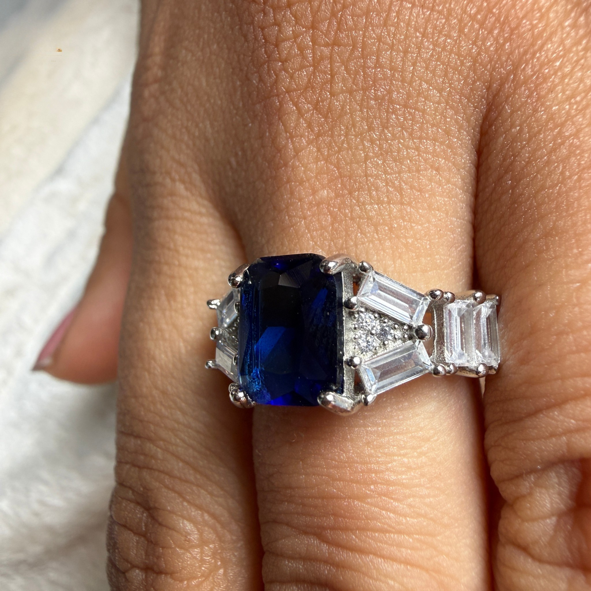 A close-up of a hand wearing a silver ring featuring a rectangular deep blue sapphire center stone and baguette-cut clear side stones.