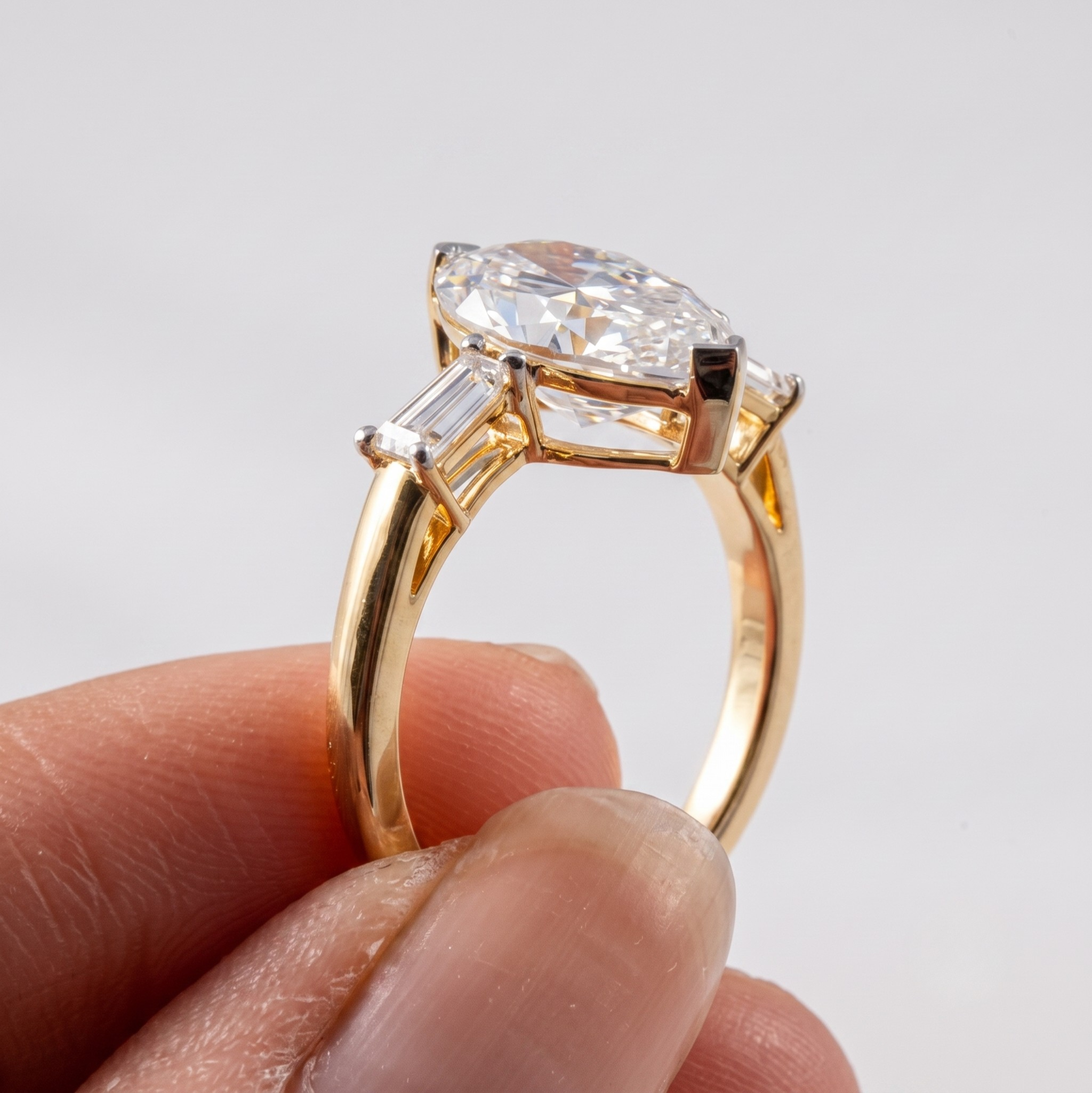 A yellow gold ring with a marquise-cut clear center stone and baguette side stones, held between fingers against a light gray background.
