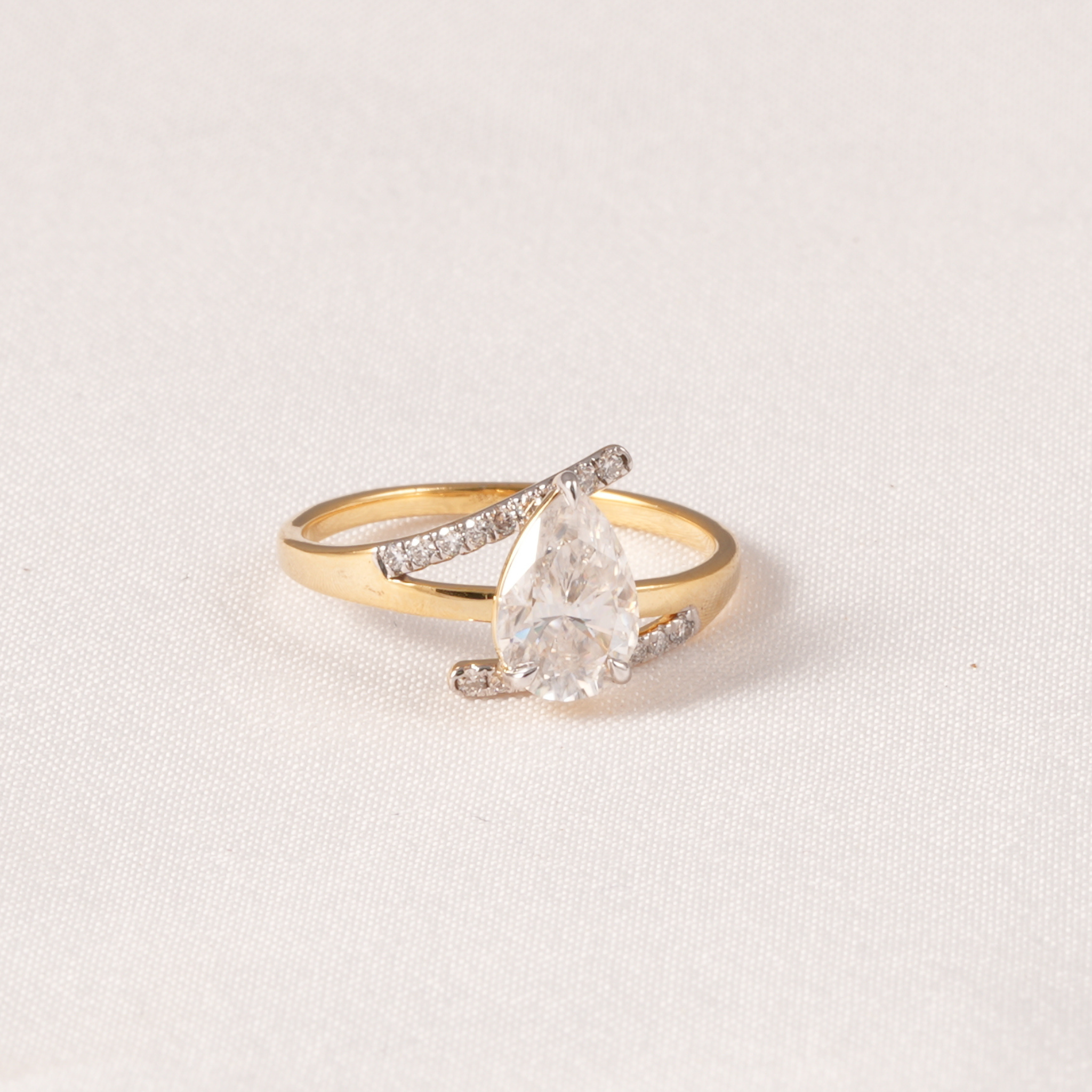 A gold ring featuring a pear-cut clear center stone and pavé-set clear accent stones on a textured off-white background.